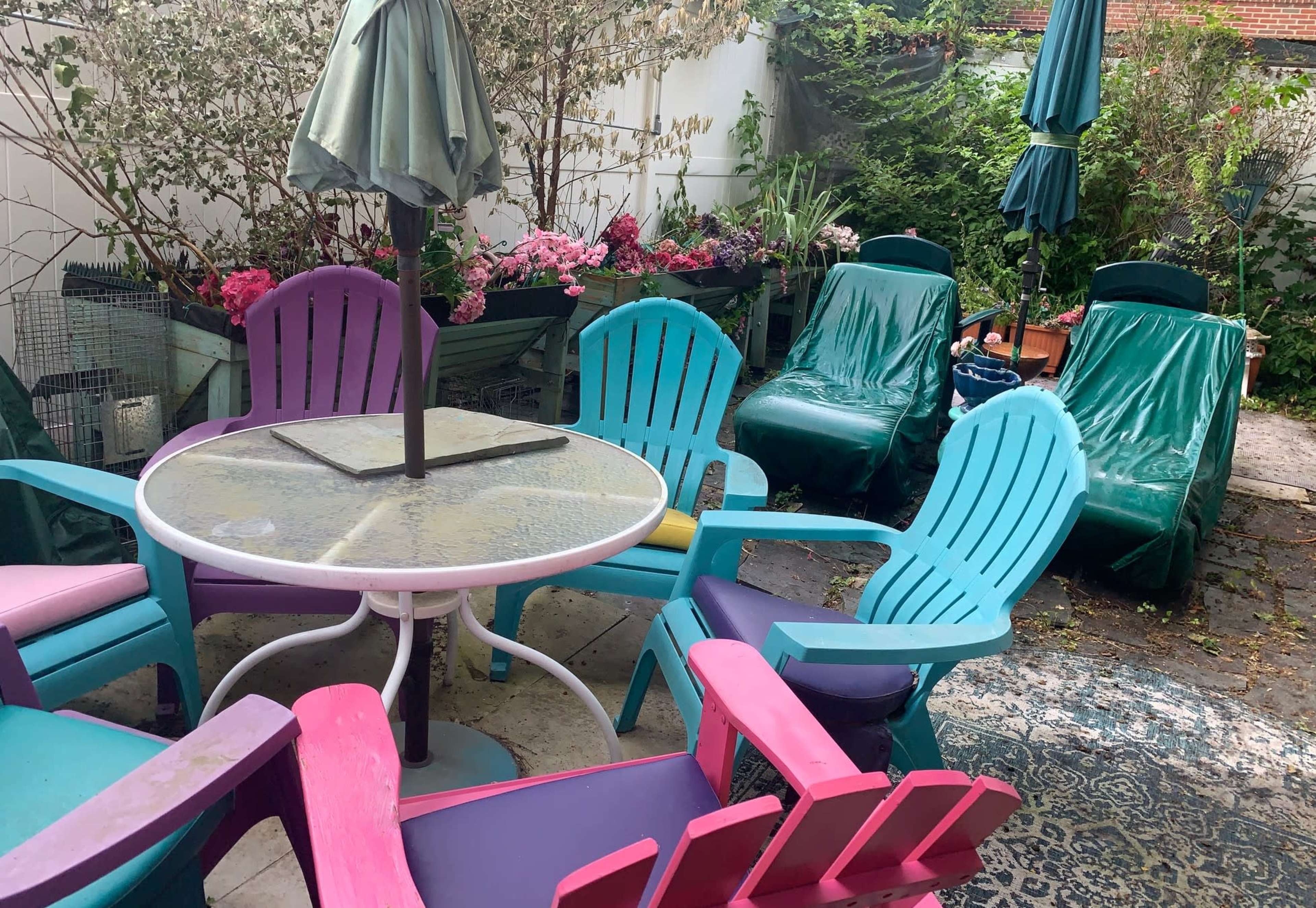 A colorful patio area features a round table surrounded by pink, blue, and purple Adirondack chairs, with lounging chairs and potted plants in the background.