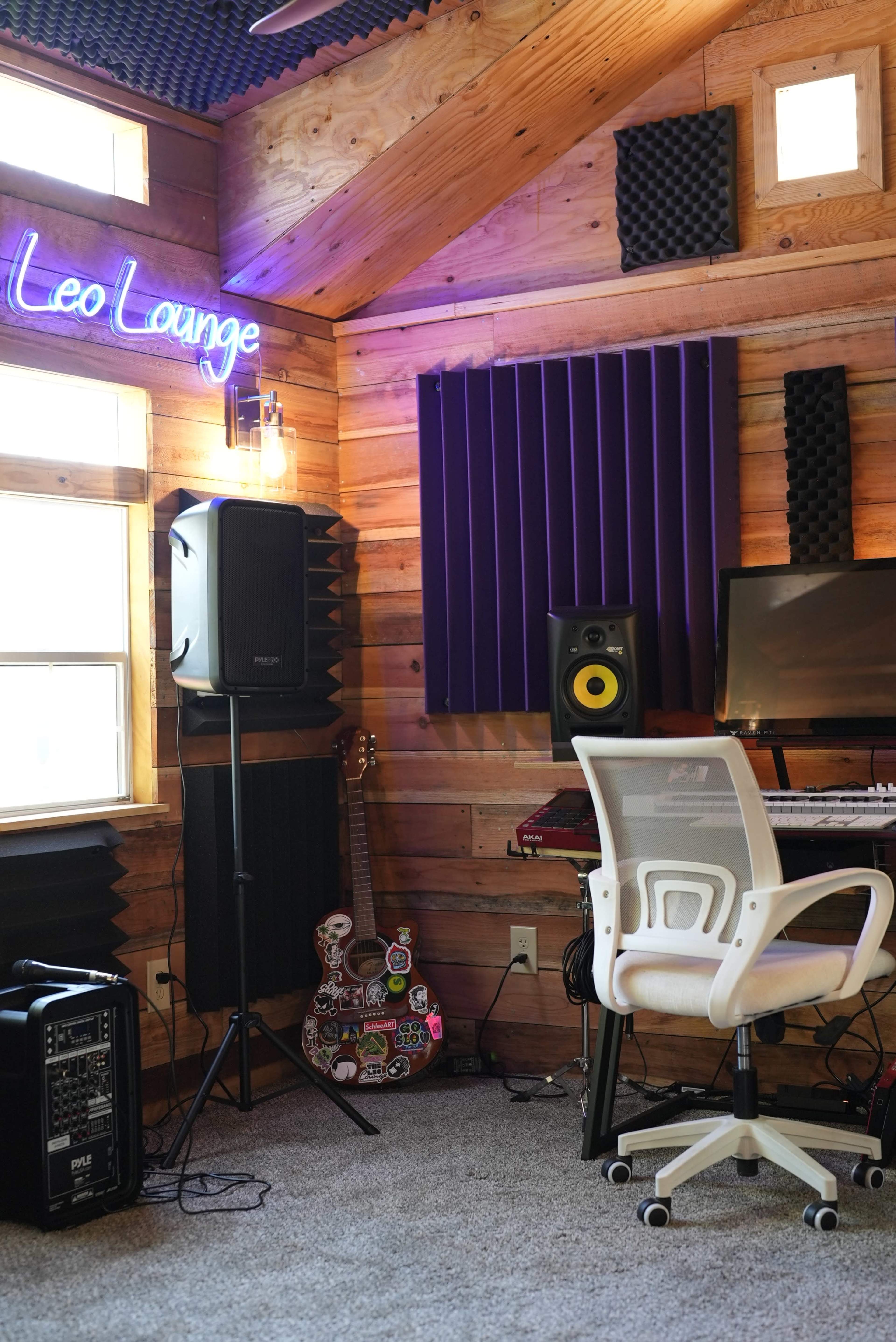 The image shows a music studio corner with wooden walls, a computer desk, speakers, a white chair, and various music equipment, including a decorated guitar.