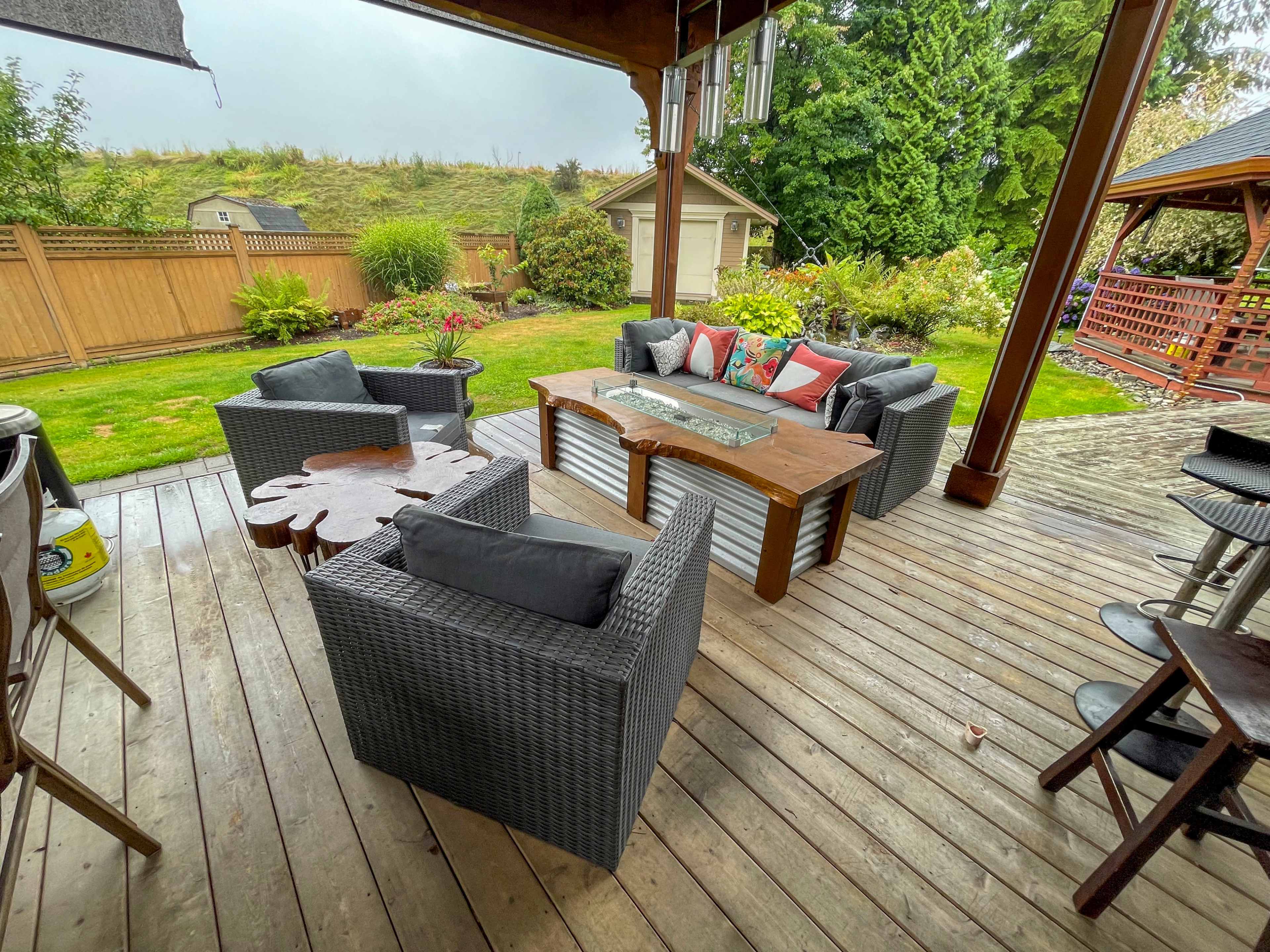 A patio area features a wooden deck with a coffee table surrounded by wicker seating and lush greenery in the background.