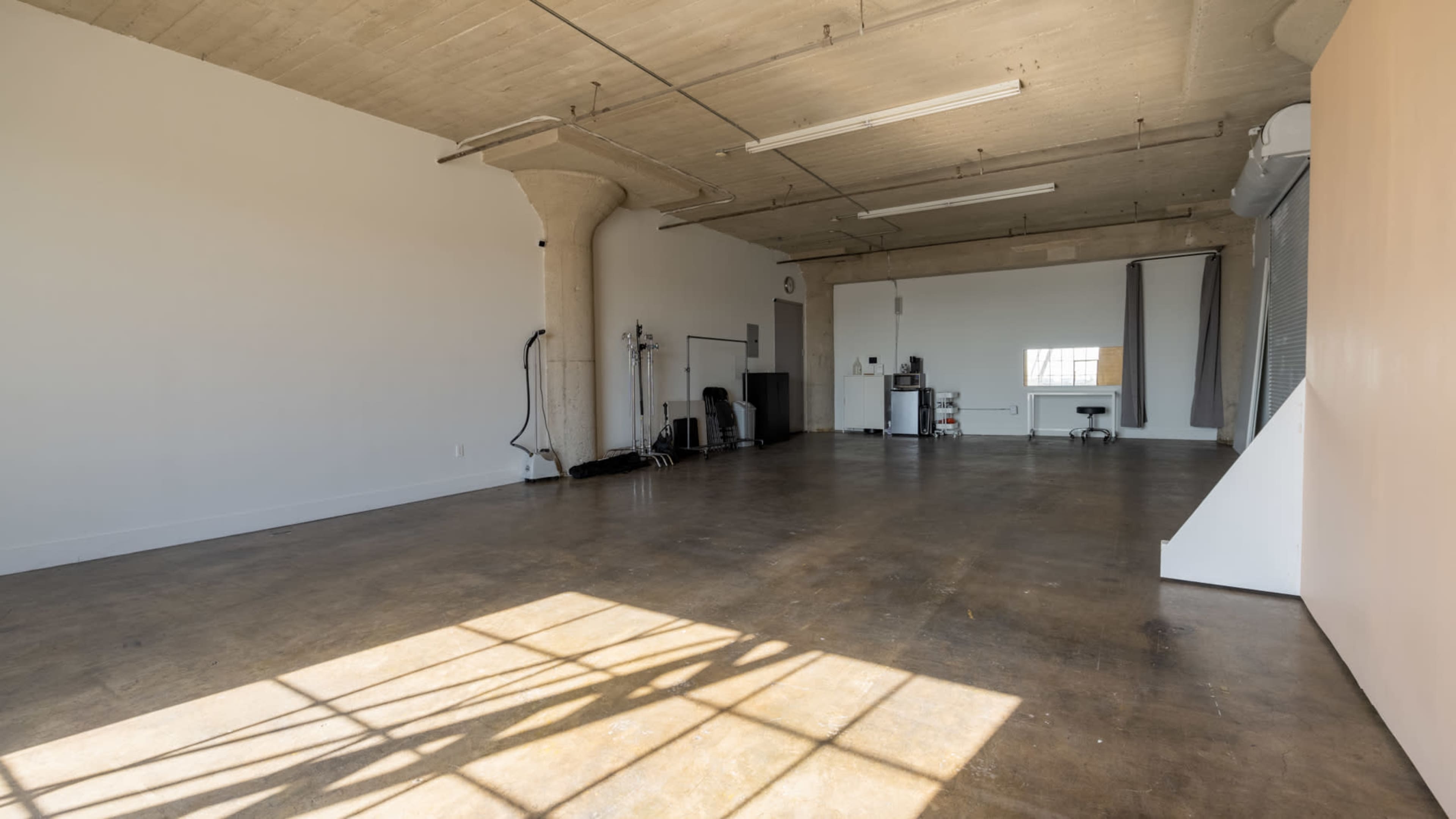 A spacious, empty room with a concrete floor, large windows, and minimal furnishings, including a small kitchenette and some workout equipment.