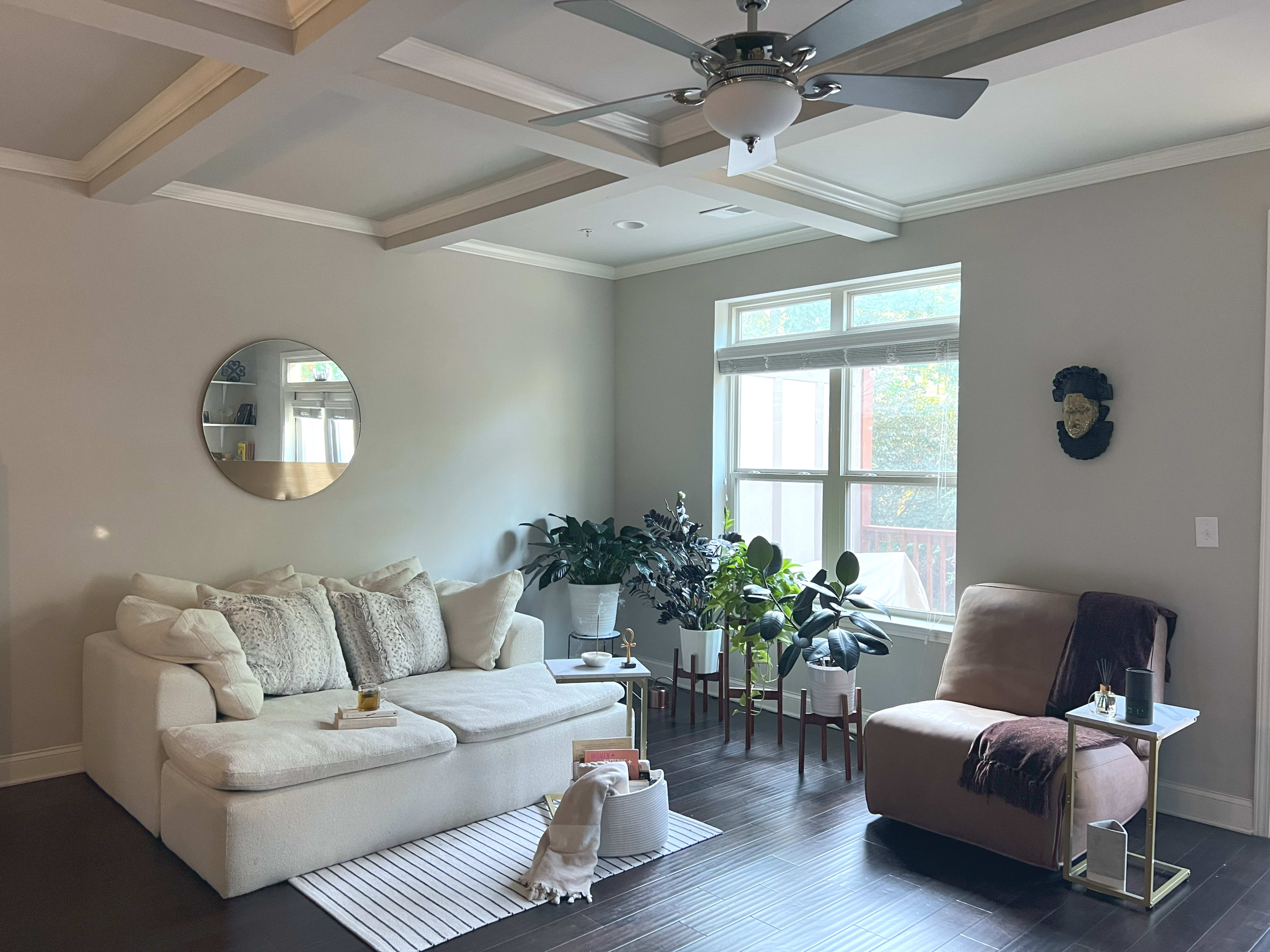 The room features a light-colored sectional sofa, a round mirror on the wall, potted plants, and a beige armchair next to a small side table.