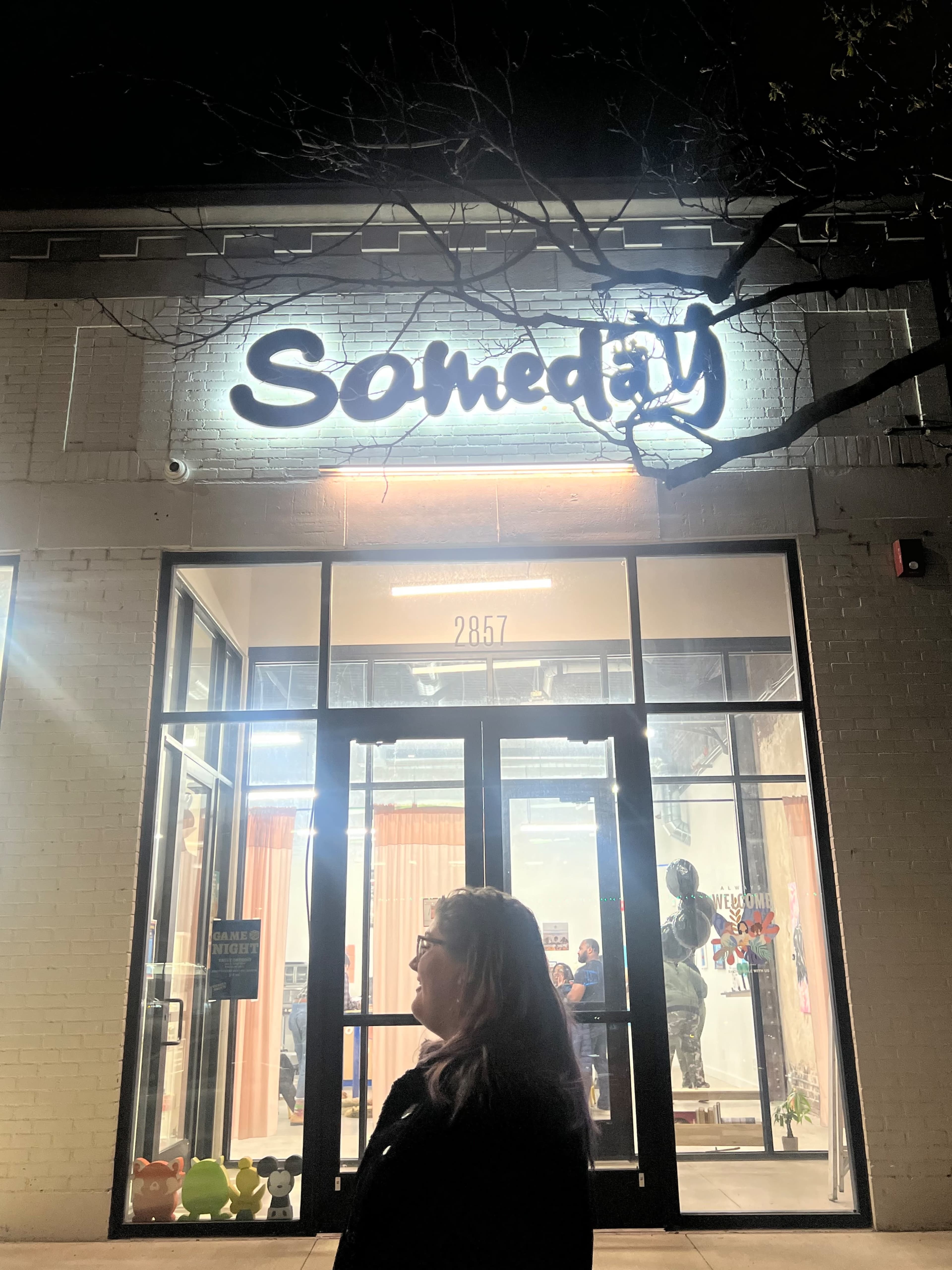 A woman stands in front of a brightly lit store called "Someday," with large windows displaying various products inside.