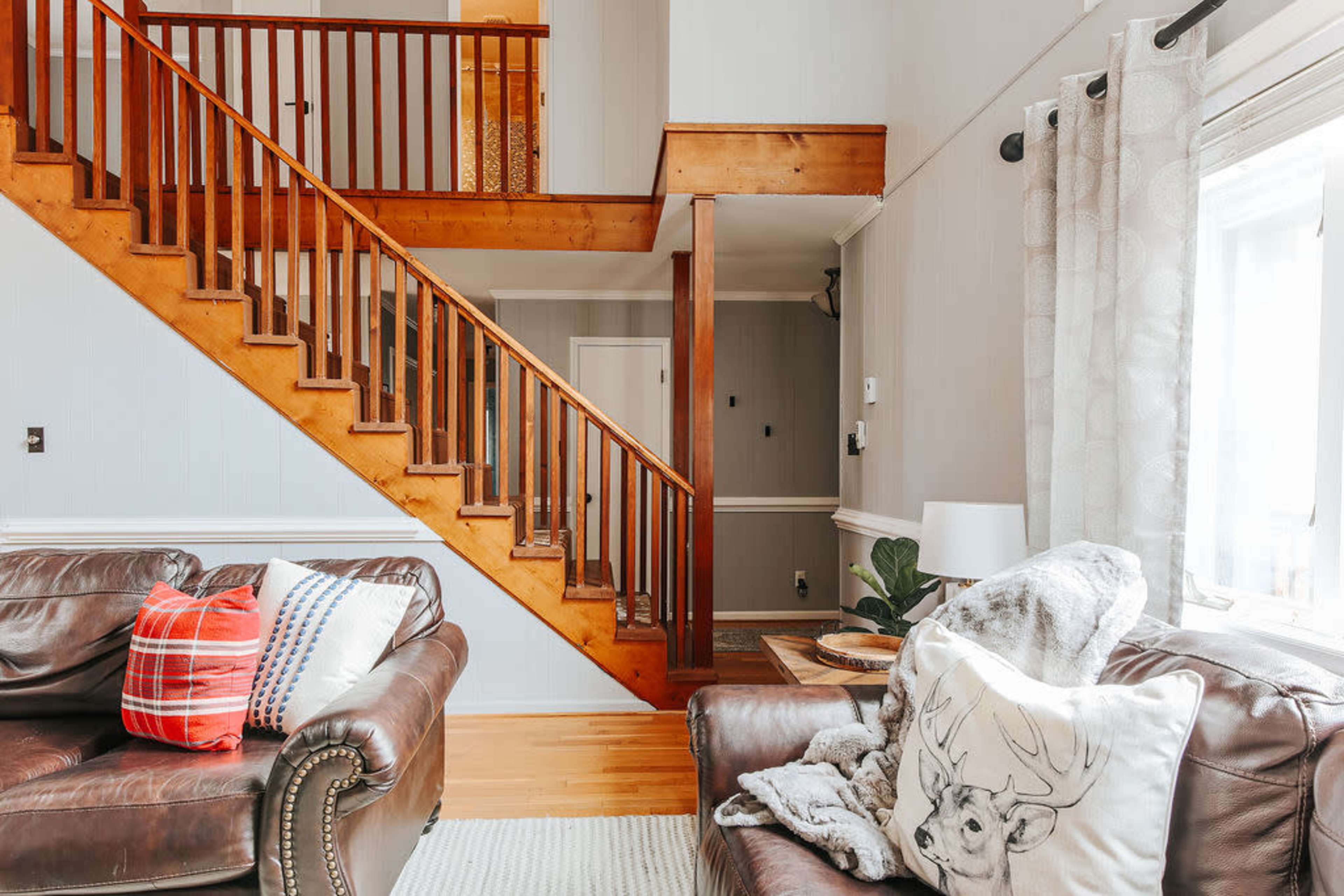 A living room with two brown leather sofas, a staircase with wooden railings, and light-colored walls adorned with curtains and decorative items.