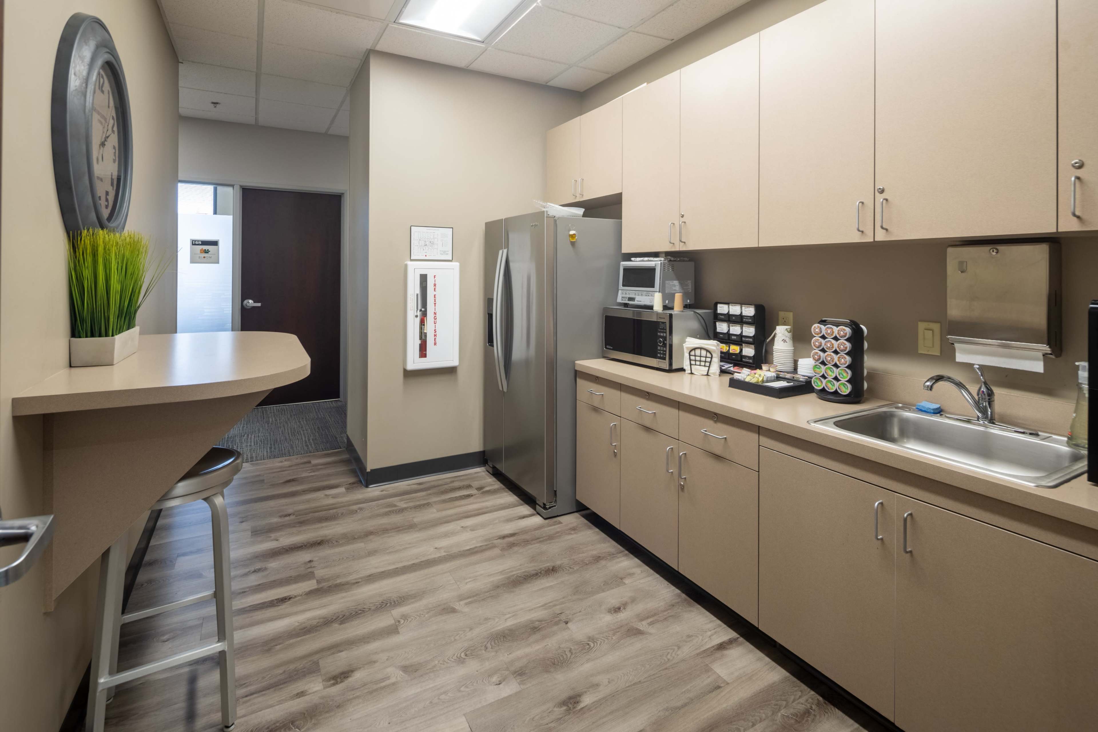 A modern break room with beige cabinets, a sink, a refrigerator, a microwave, and a coffee station, along with a small bar area with a stool.