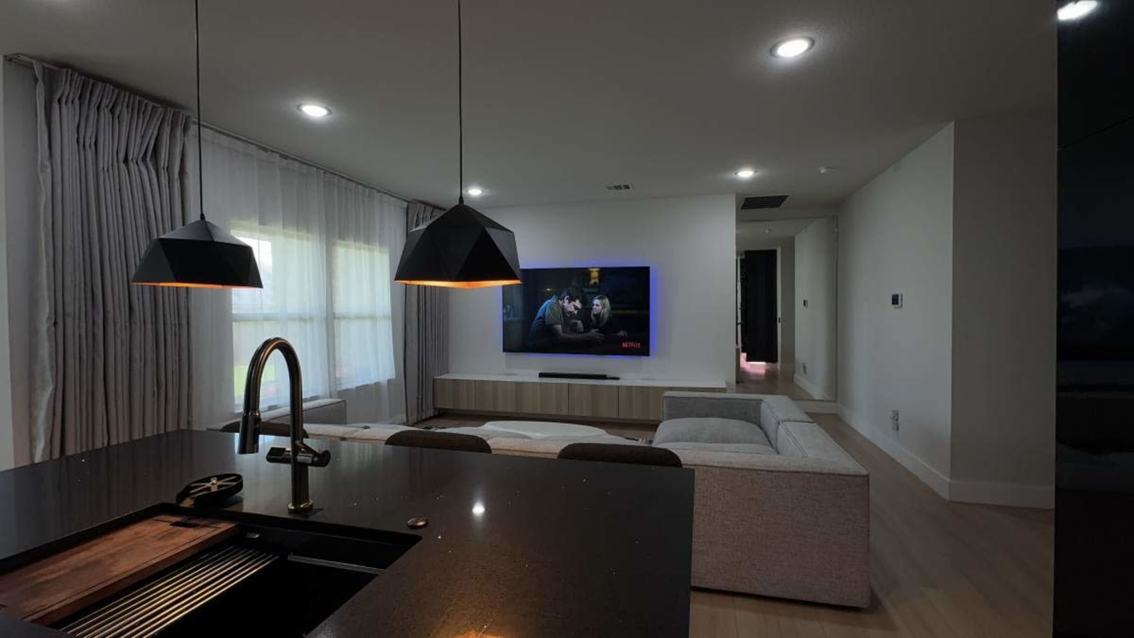 A modern living room features a couch facing a mounted television, with pendant lights hanging over a kitchen island in the foreground.