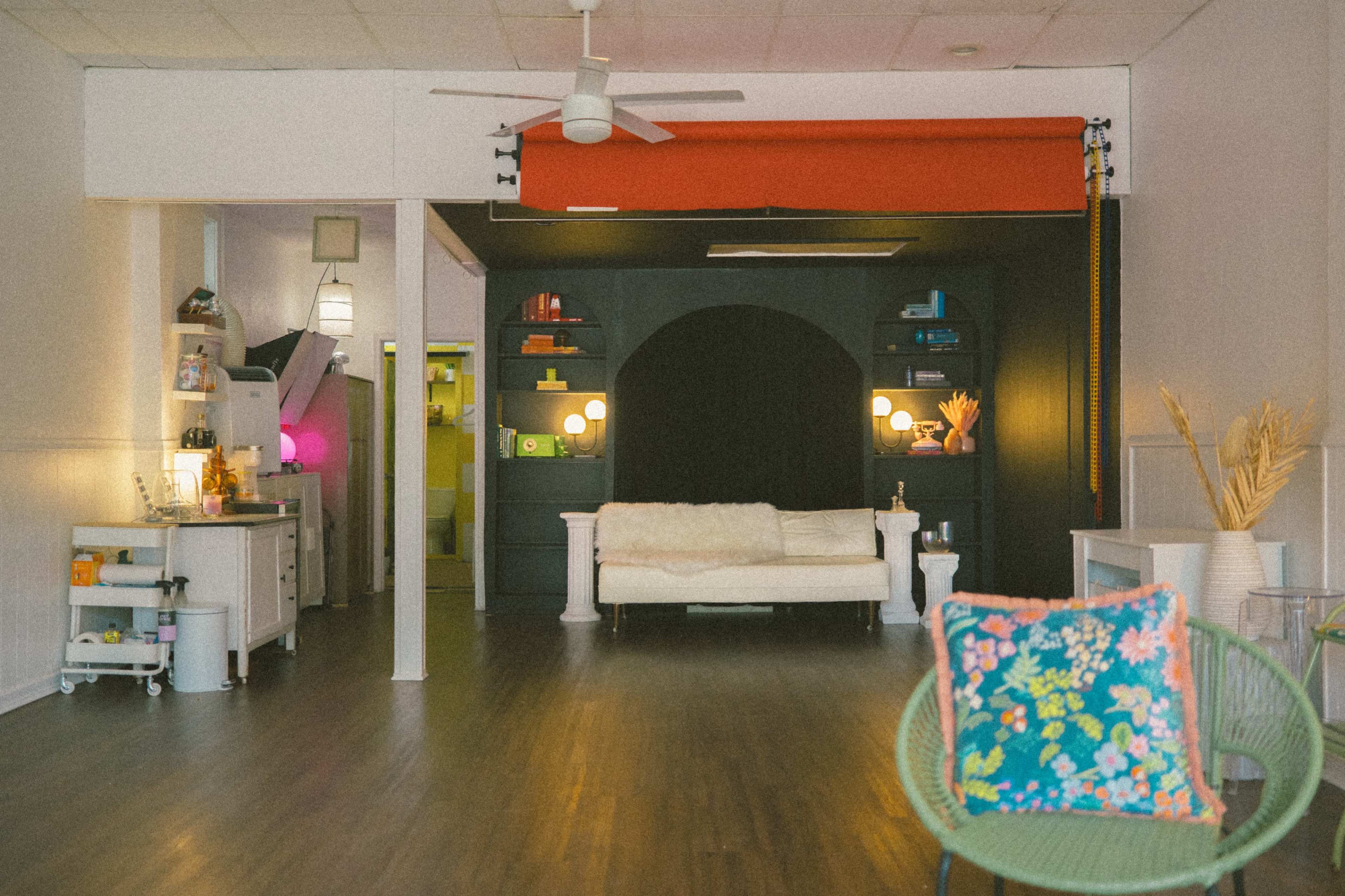 A spacious room featuring a white couch, black shelves with decorative items, and a vibrant red curtain, all complemented by wooden flooring and modern lighting.
