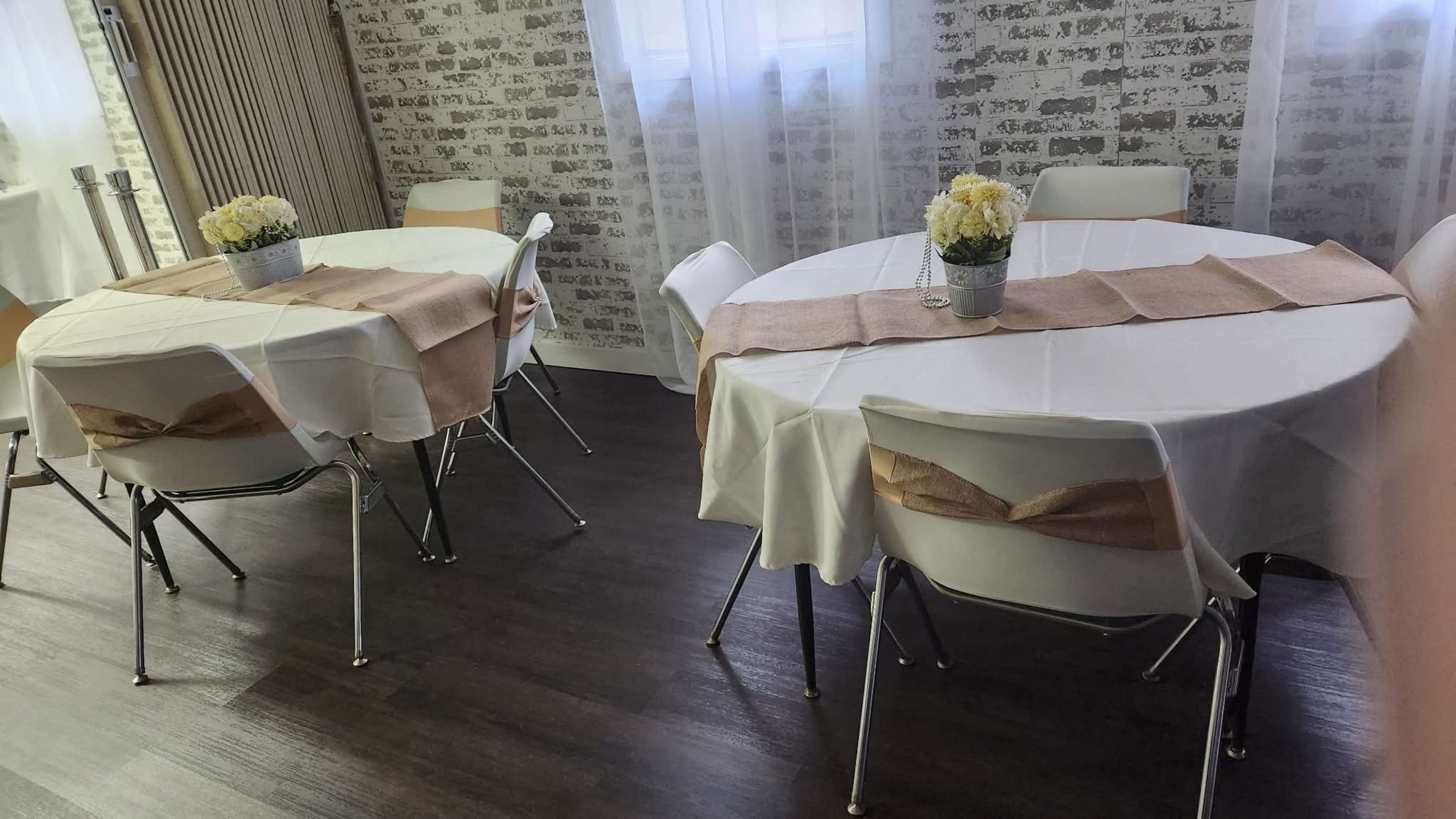 Two round tables are set with white tablecloths and centerpieces of flowers in a room with a brick wall and large windows.