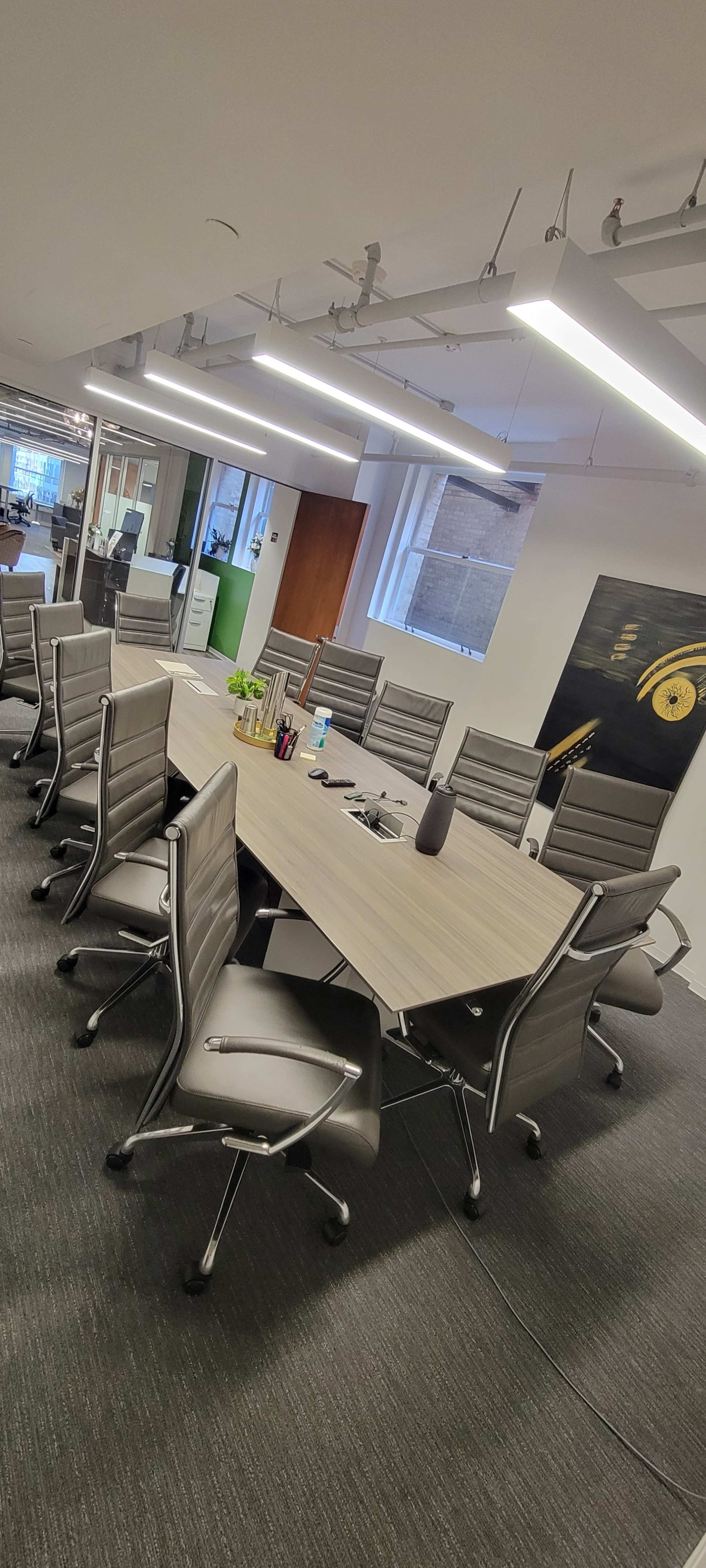 A long conference table surrounded by ergonomic chairs is positioned in a modern office meeting room with large windows and minimal decor.