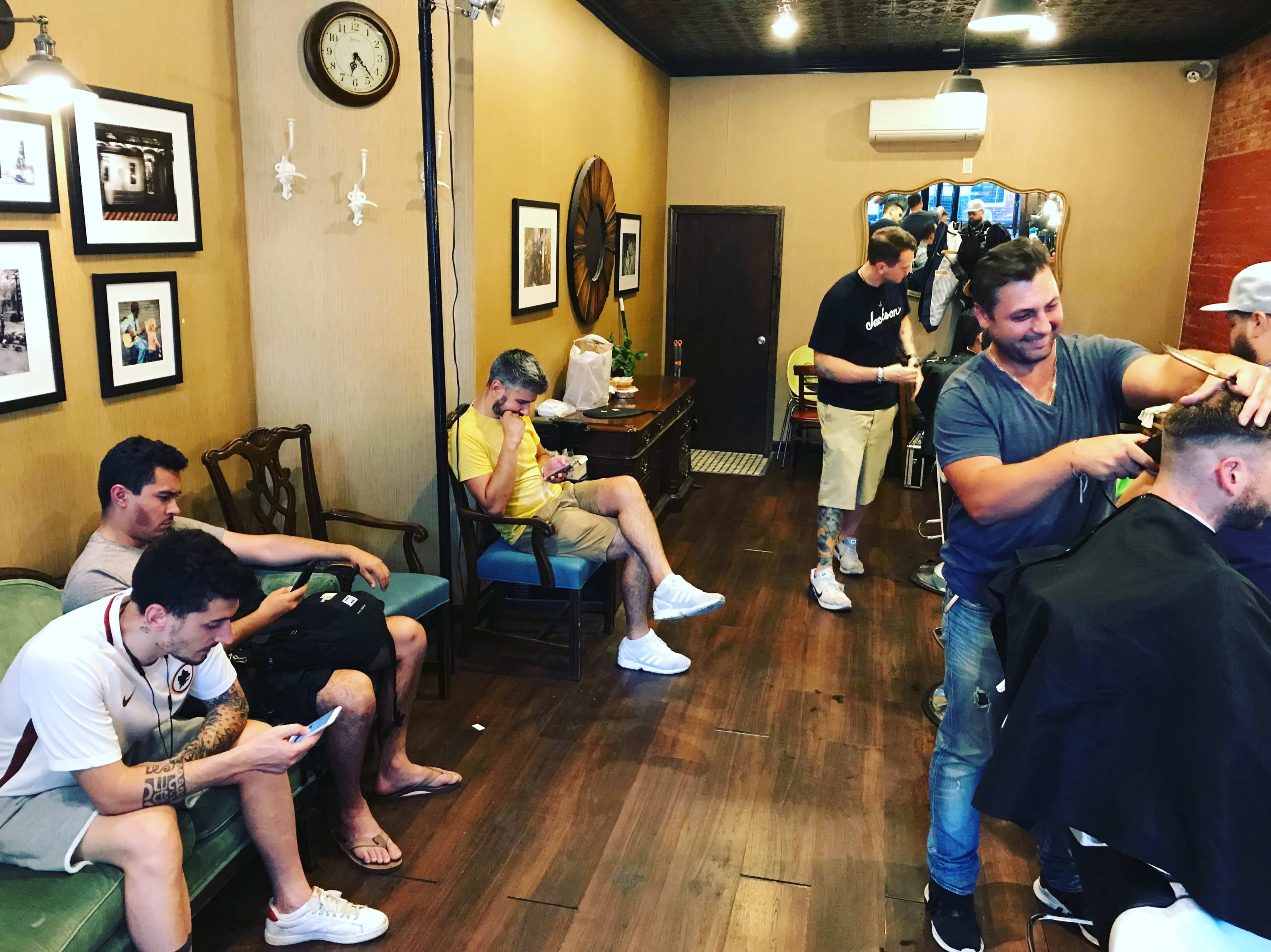 A barbershop features several customers waiting and being served, with chairs arranged along the wall and barbers attending to clients.