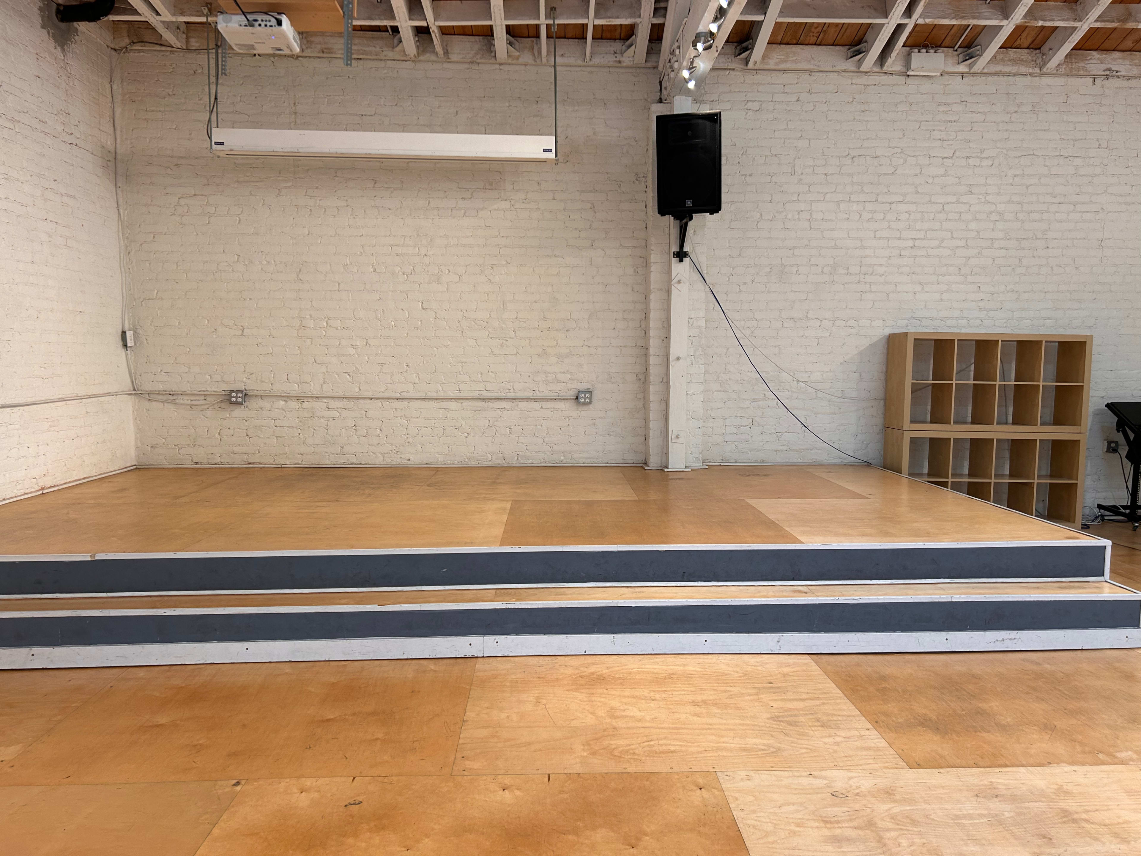 A small stage with three steps, surrounded by a plain white wall and wooden flooring.
