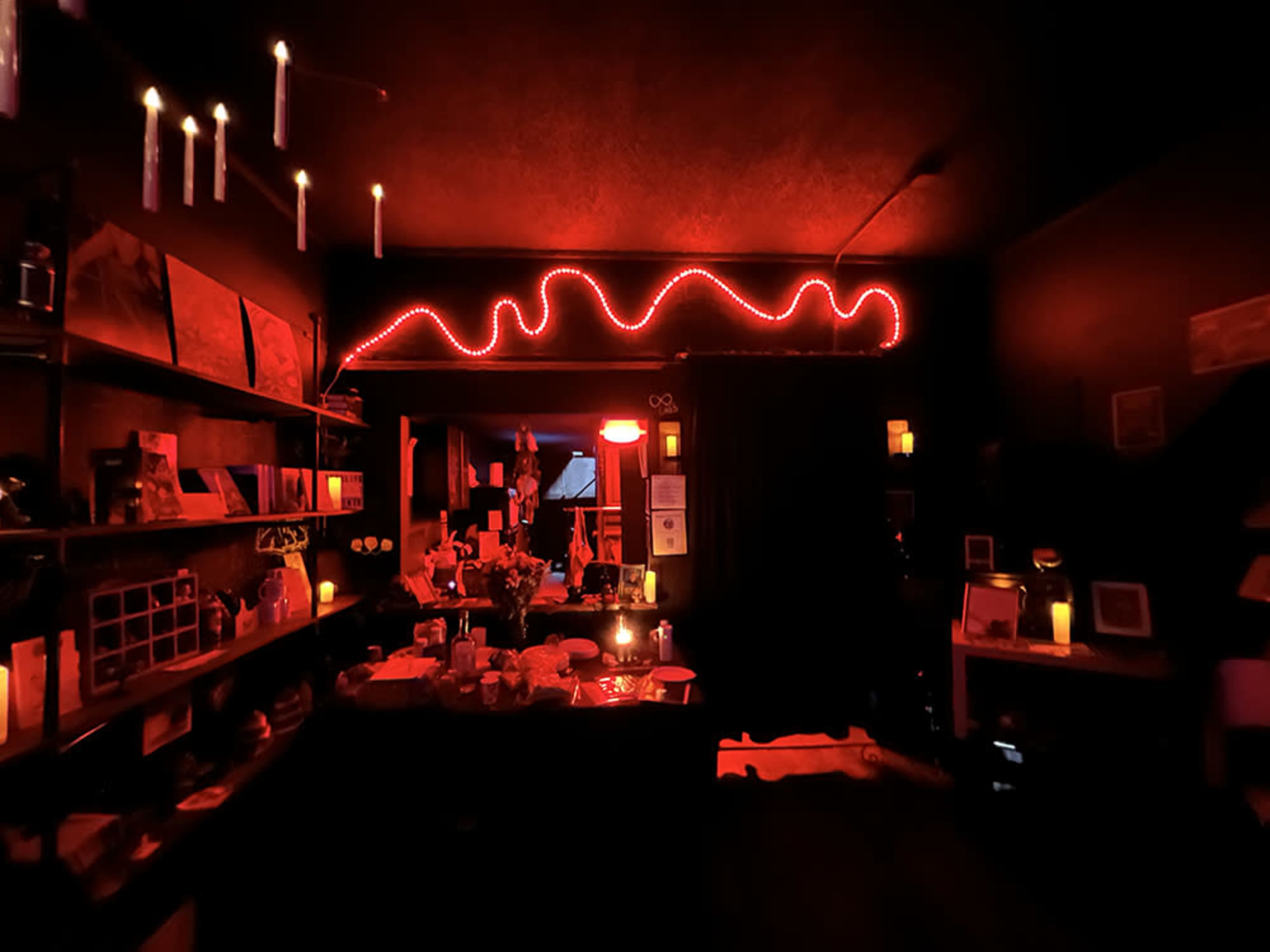 A dimly lit room featuring a red neon light, shelves filled with various items, and several candles scattered throughout the space.