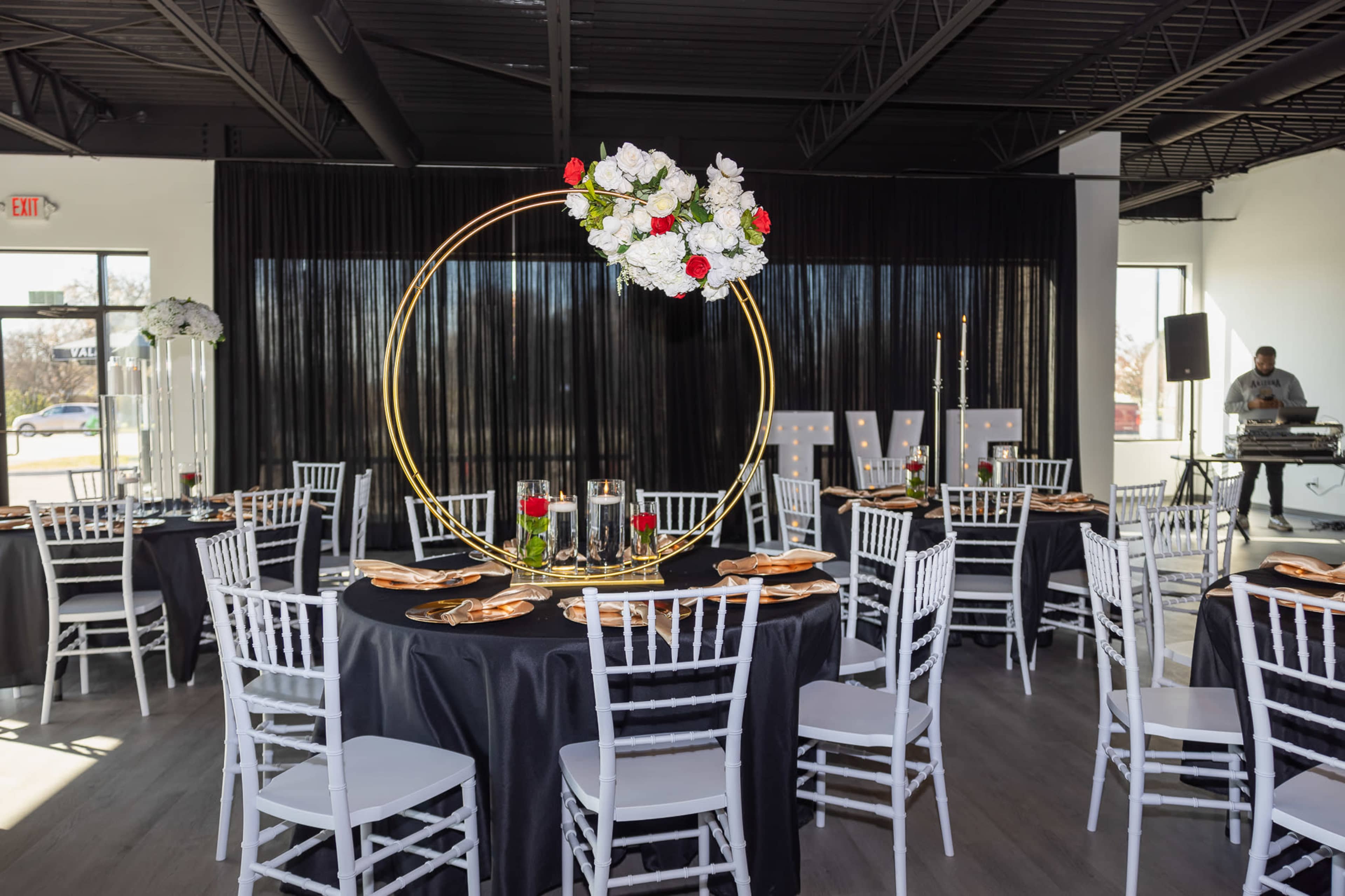 A banquet hall is set up for an event, featuring round tables with black tablecloths, white chiavari chairs, and a large floral arrangement in a gold ring as a centerpiece.