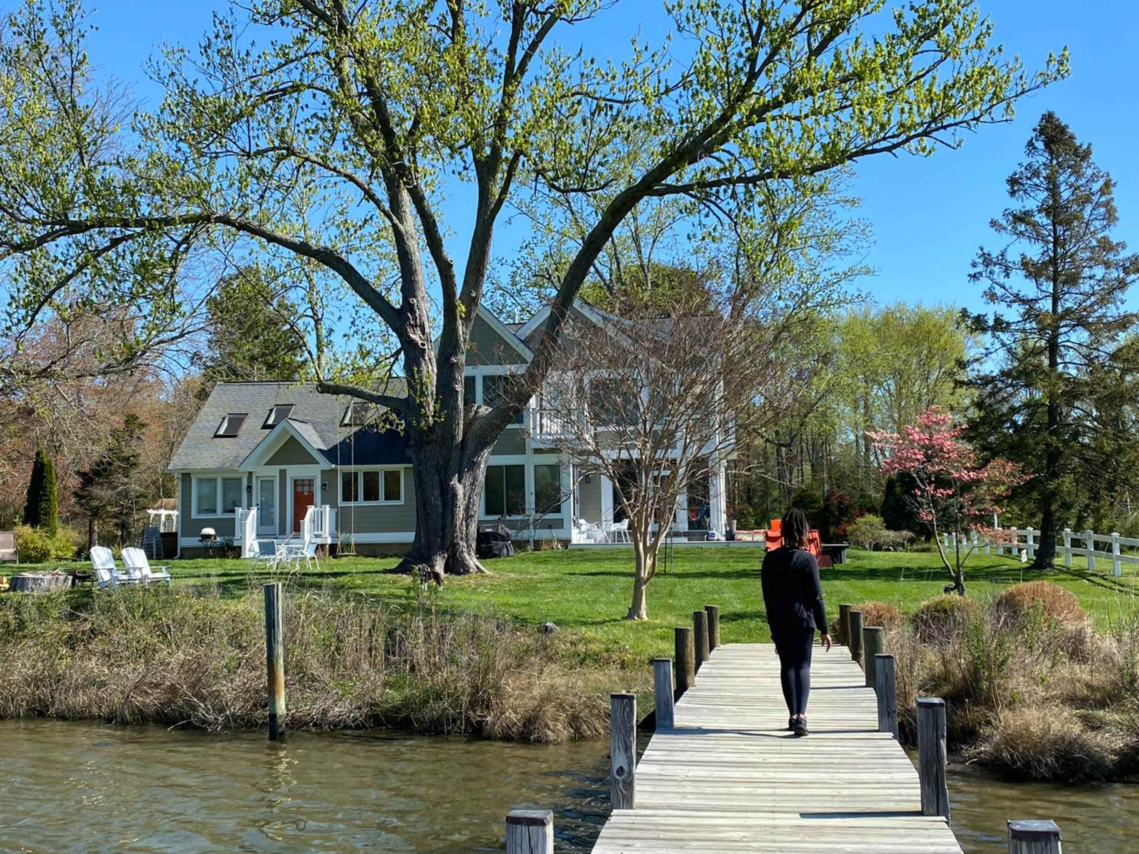 A person walks along a wooden dock towards a house near a tree-lined waterfront.