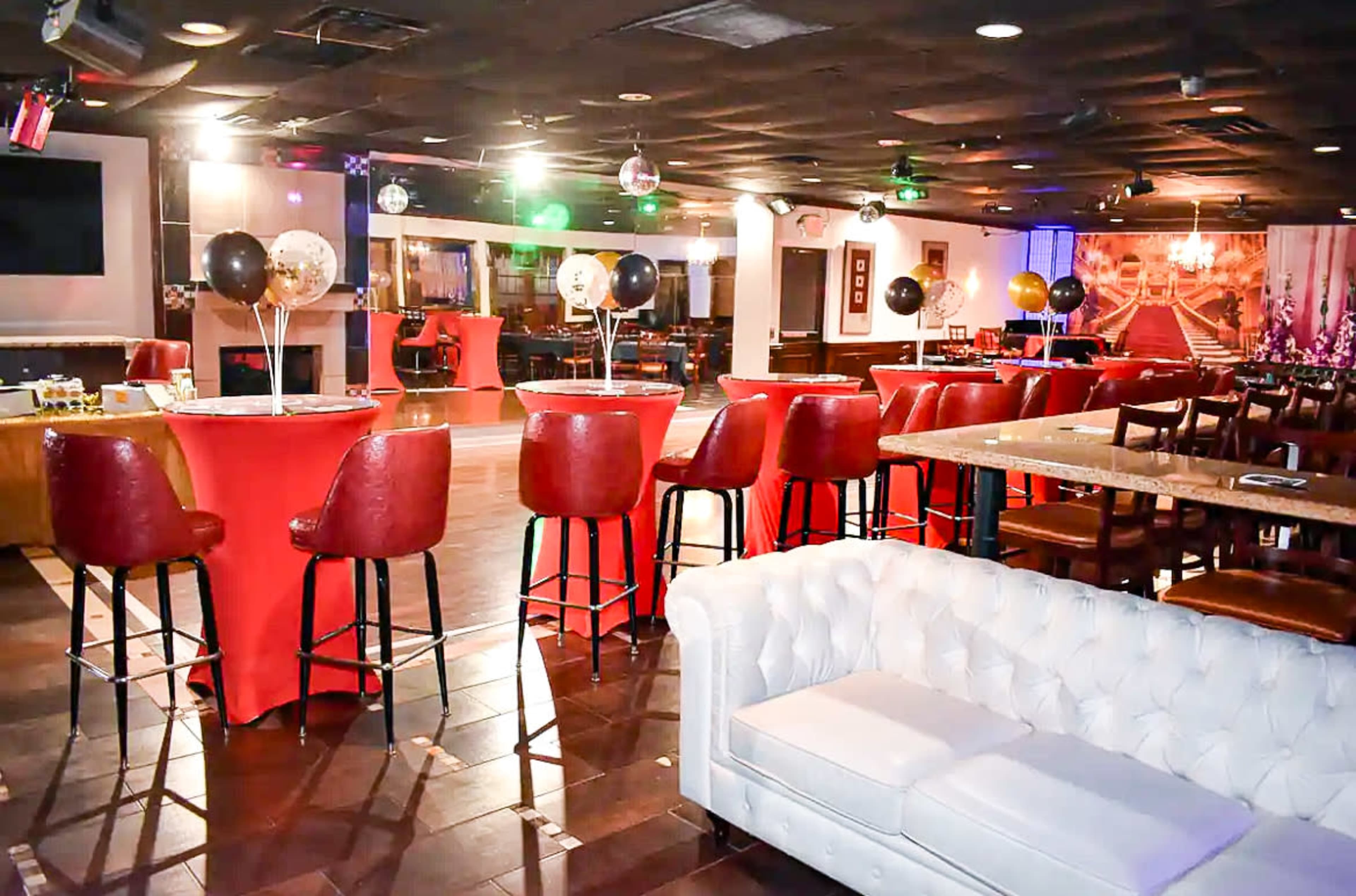 The interior of a lounge features red bar stools and tables, a white couch, and colorful lights illuminating the space.