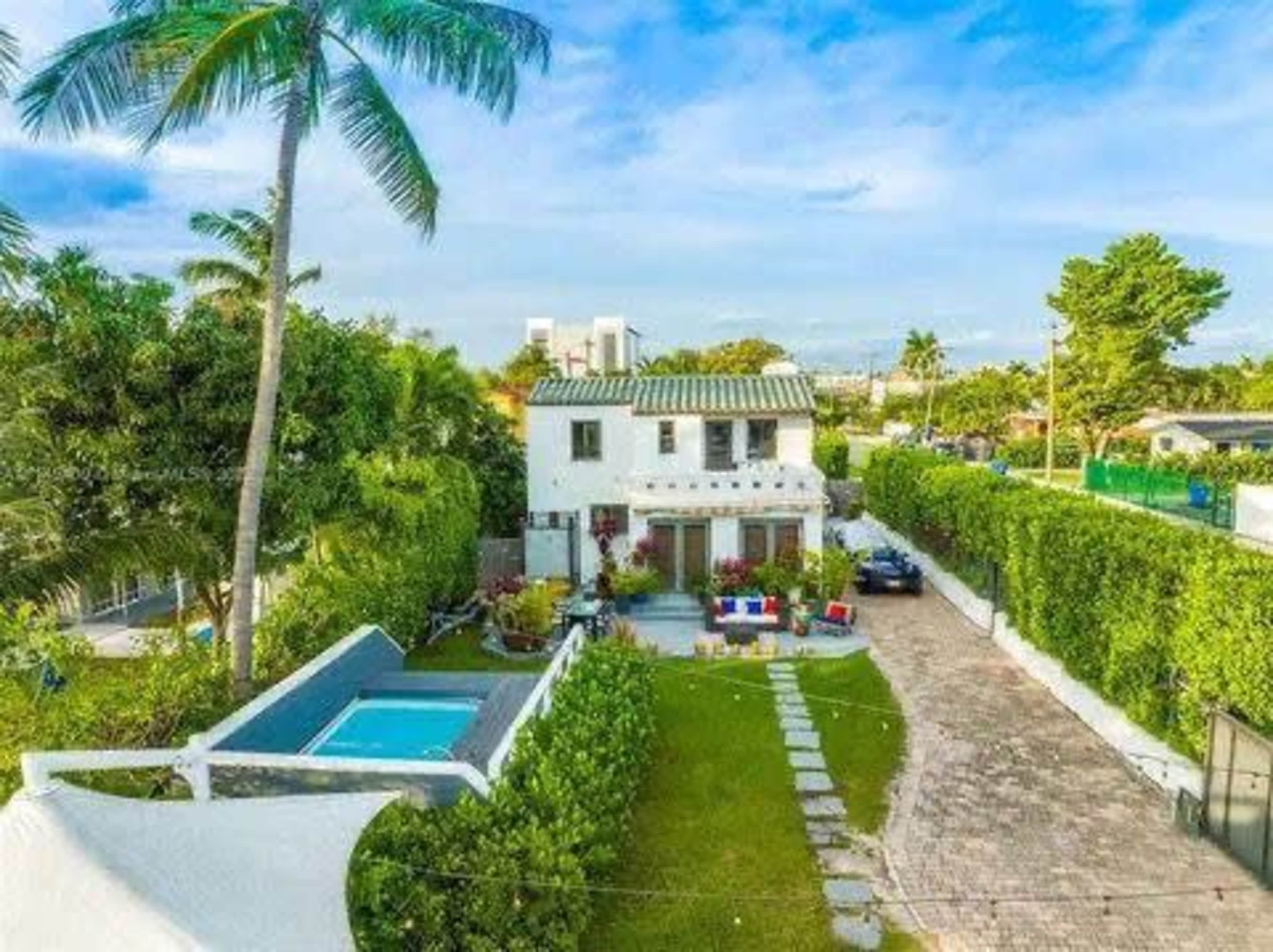 The image shows a two-story white house surrounded by greenery, with a swimming pool and a pathway leading to it.