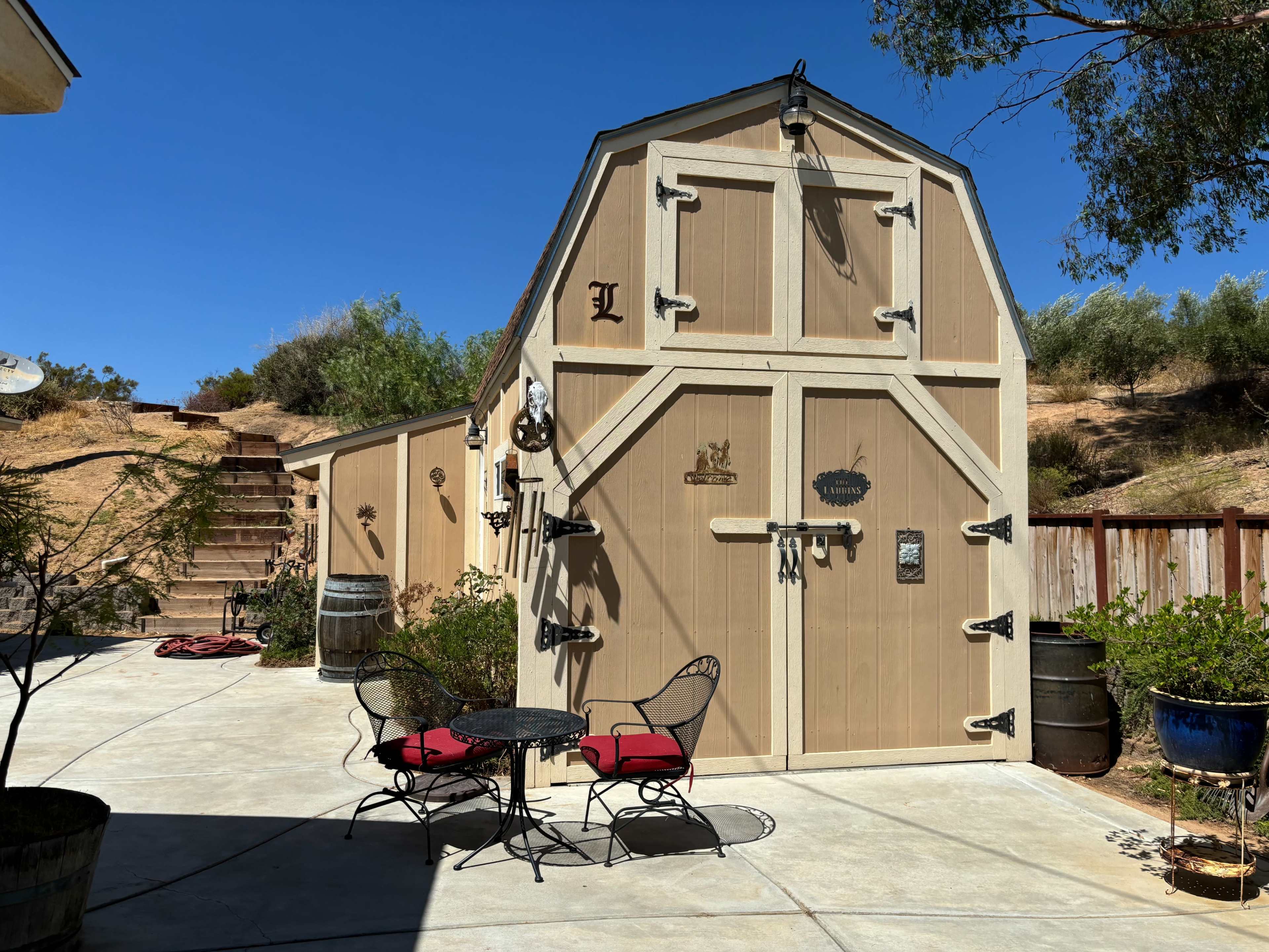A two-story shed with a pitched roof and various decorative items stands in a concrete patio area surrounded by greenery.