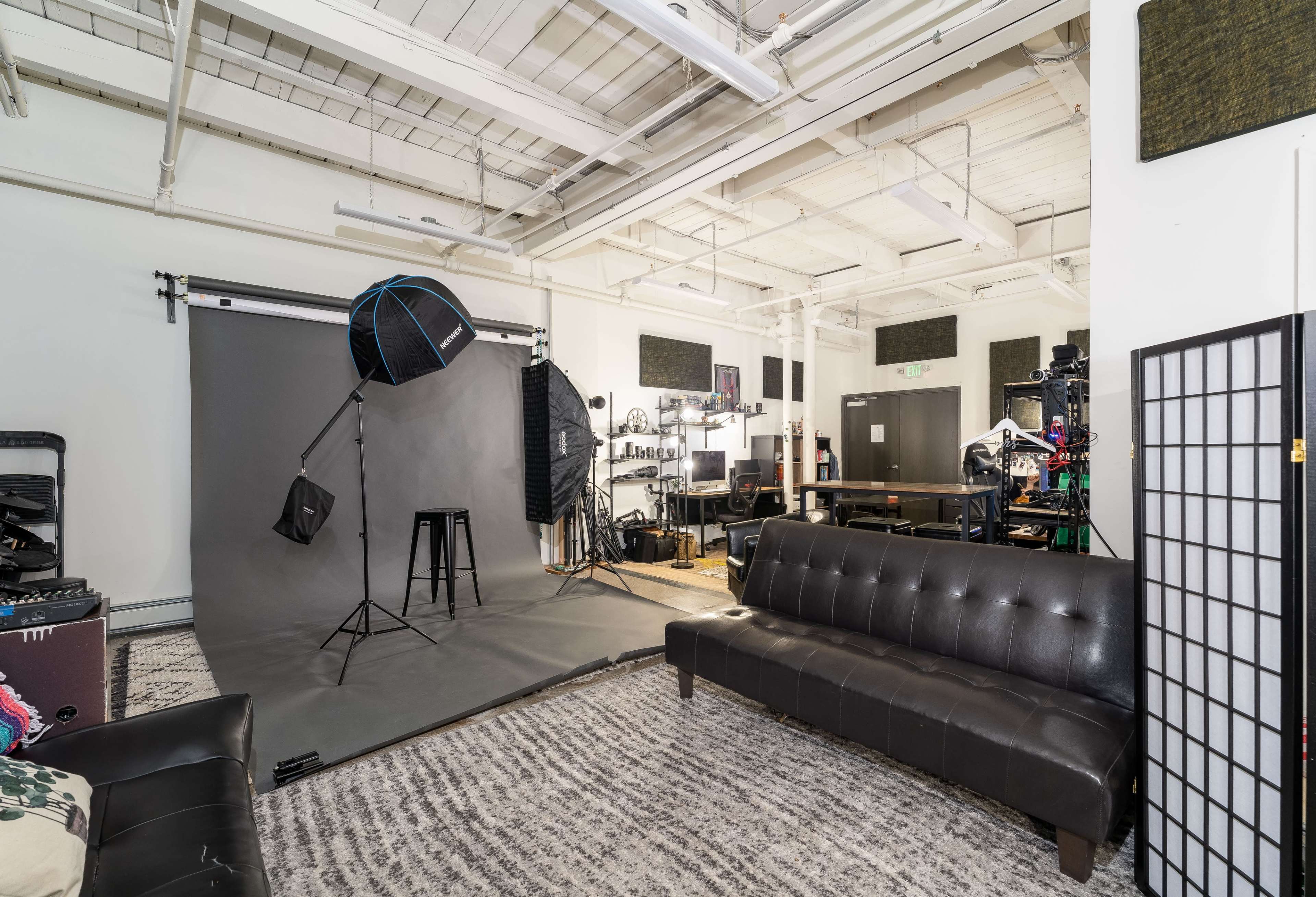 A photography studio featuring a gray backdrop, a light stand with an umbrella, a black stool, and a seating area with a dark sofa.