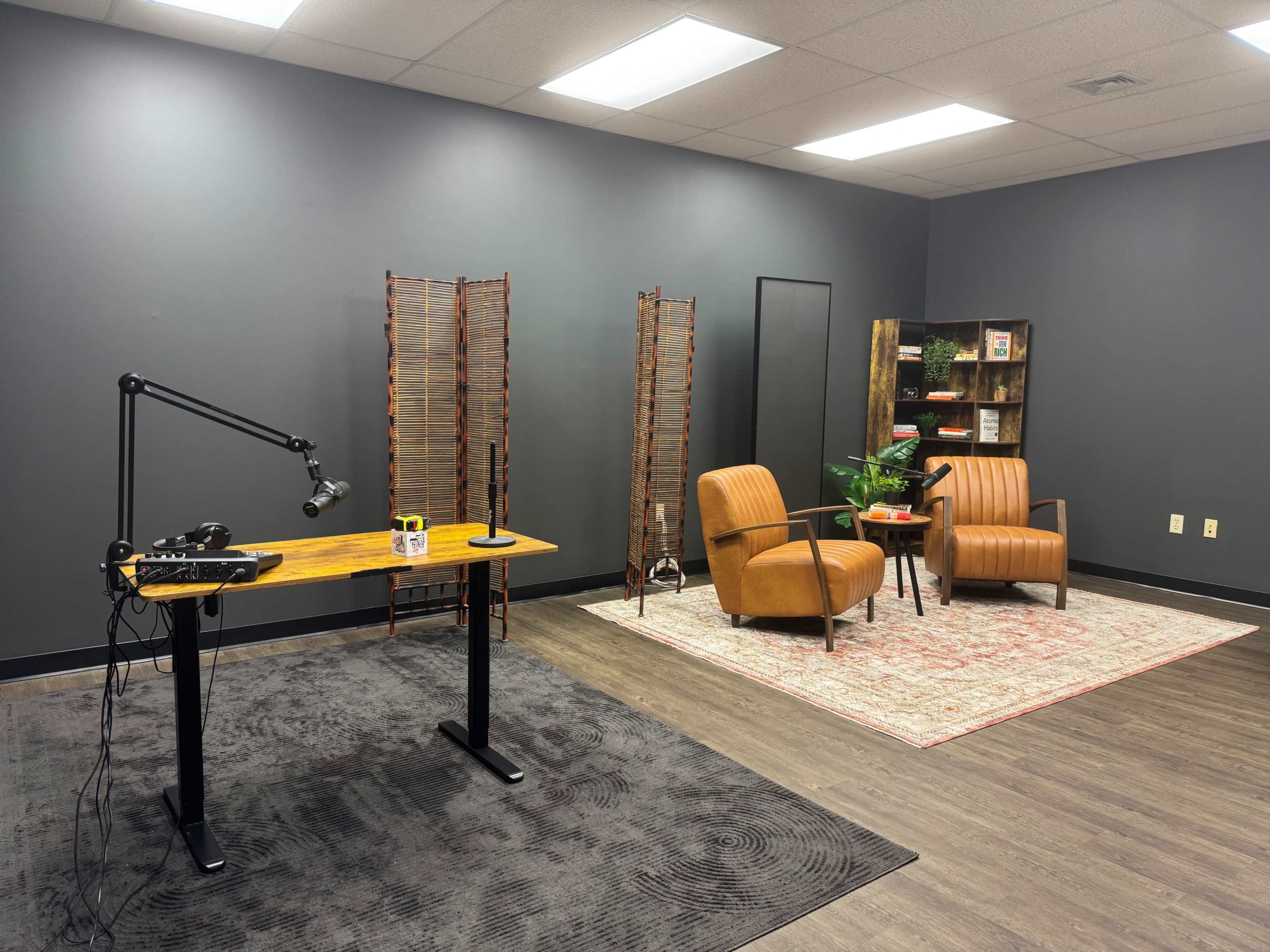 The image shows a modern office space featuring a wooden desk with a microphone set up, two orange chairs, a decorative screen, and a bookshelf against a gray wall.