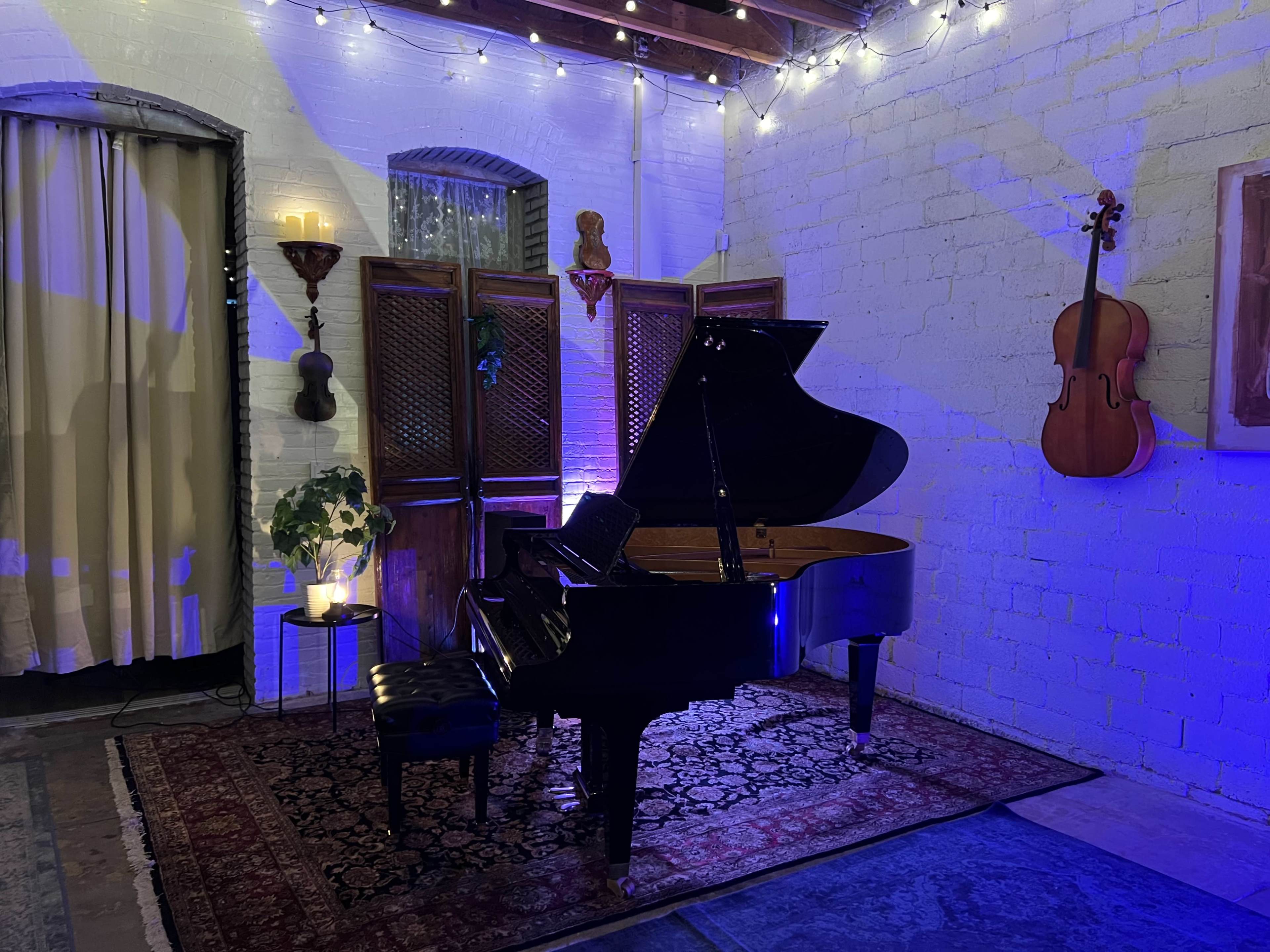 A grand piano sits on a patterned rug under soft, blue lighting, next to a wooden screen and a wall-mounted cello in a cozy room.