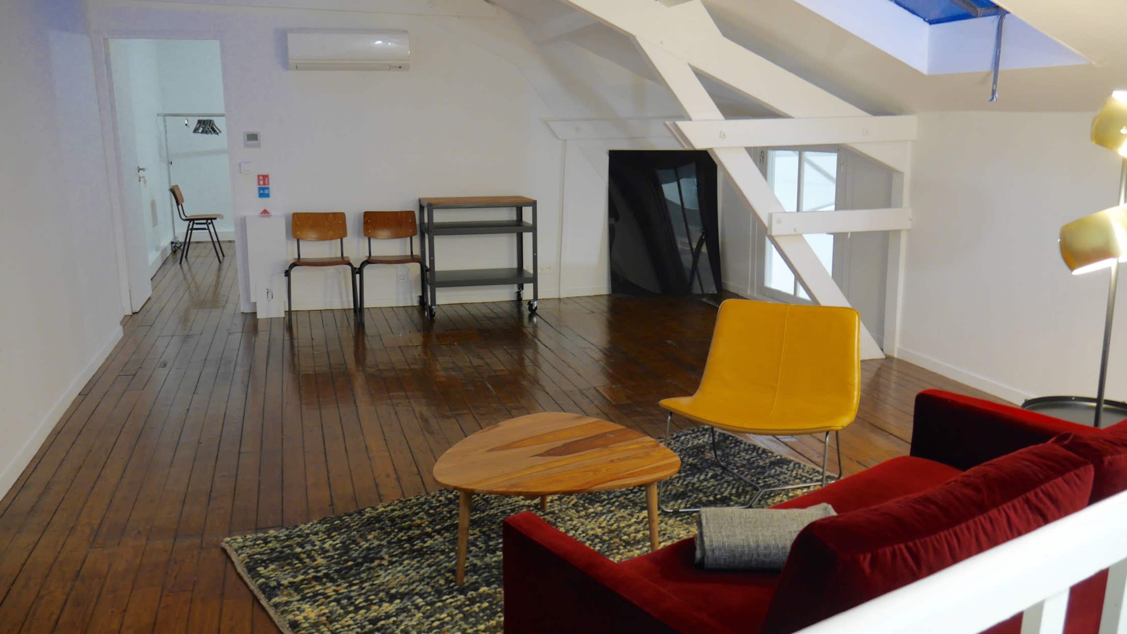 The image shows a spacious, bright room with wooden floors, featuring a red sofa, a yellow chair, a round wooden table, a shelf, and three wooden chairs.