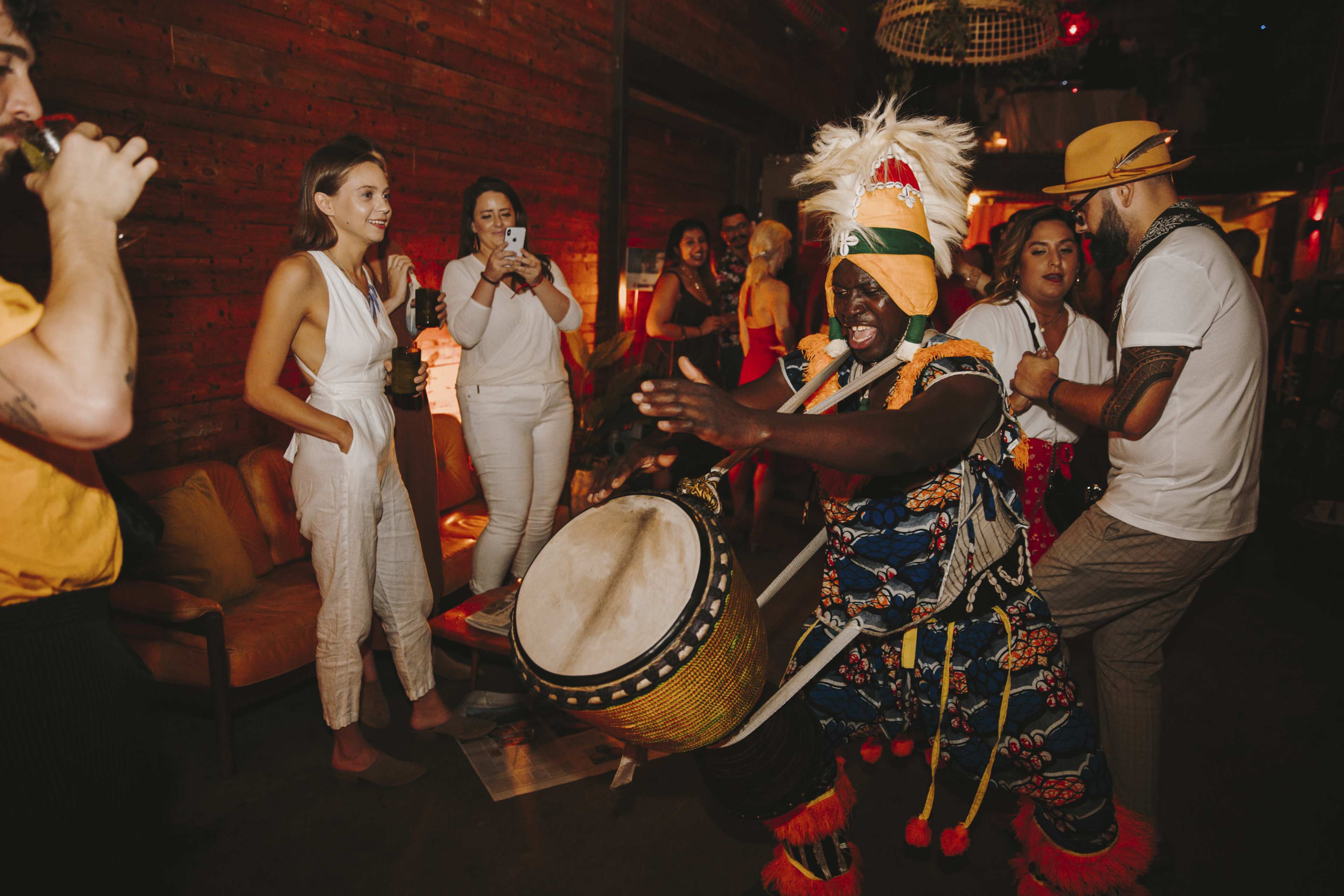 A group of people gathers in a dimly lit venue as a drummer performs energetically, surrounded by onlookers who are capturing the moment with their phones.