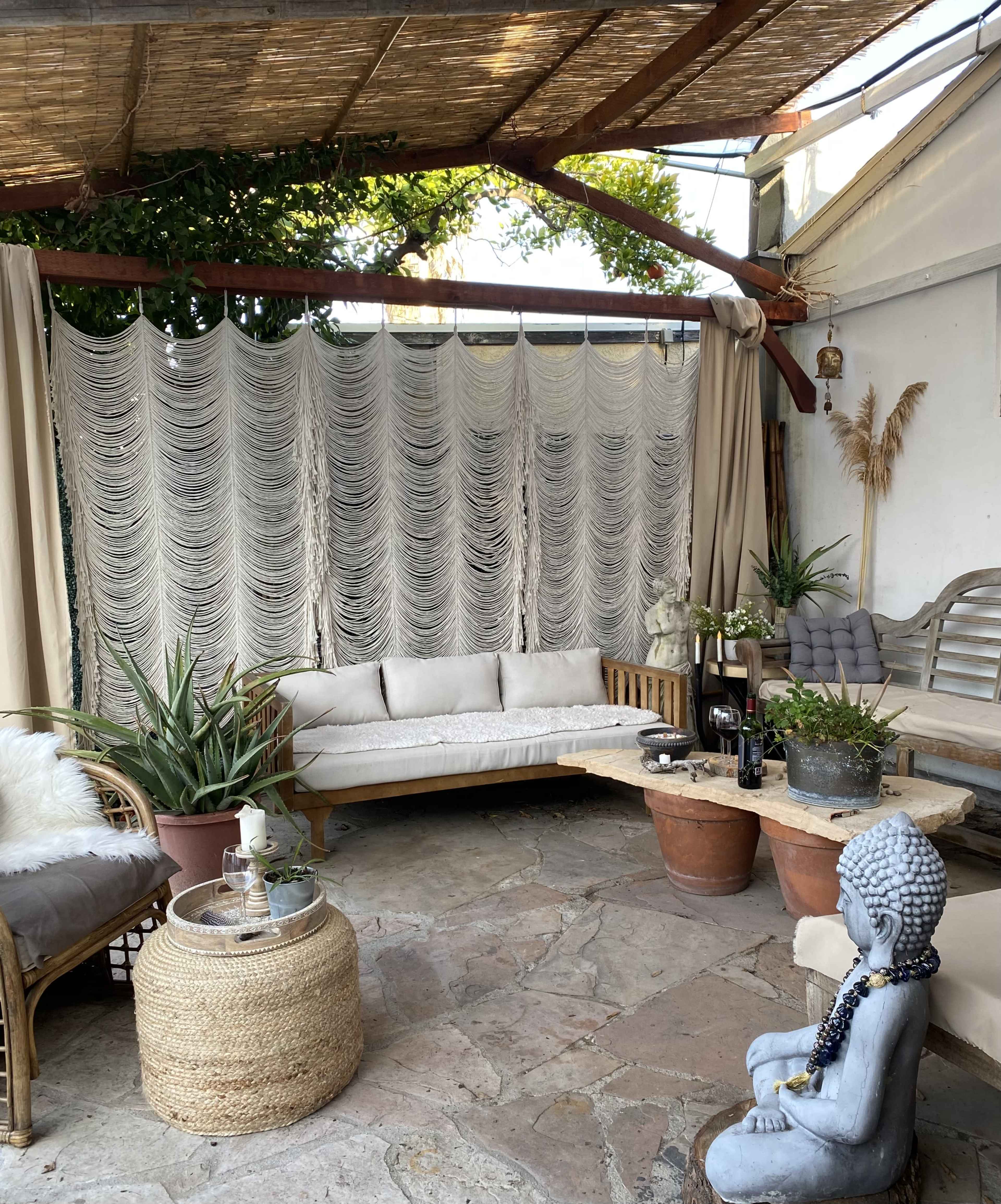 The scene features a cozy outdoor seating area with a light-colored sofa, a wooden coffee table, potted plants, and a decorative statue.