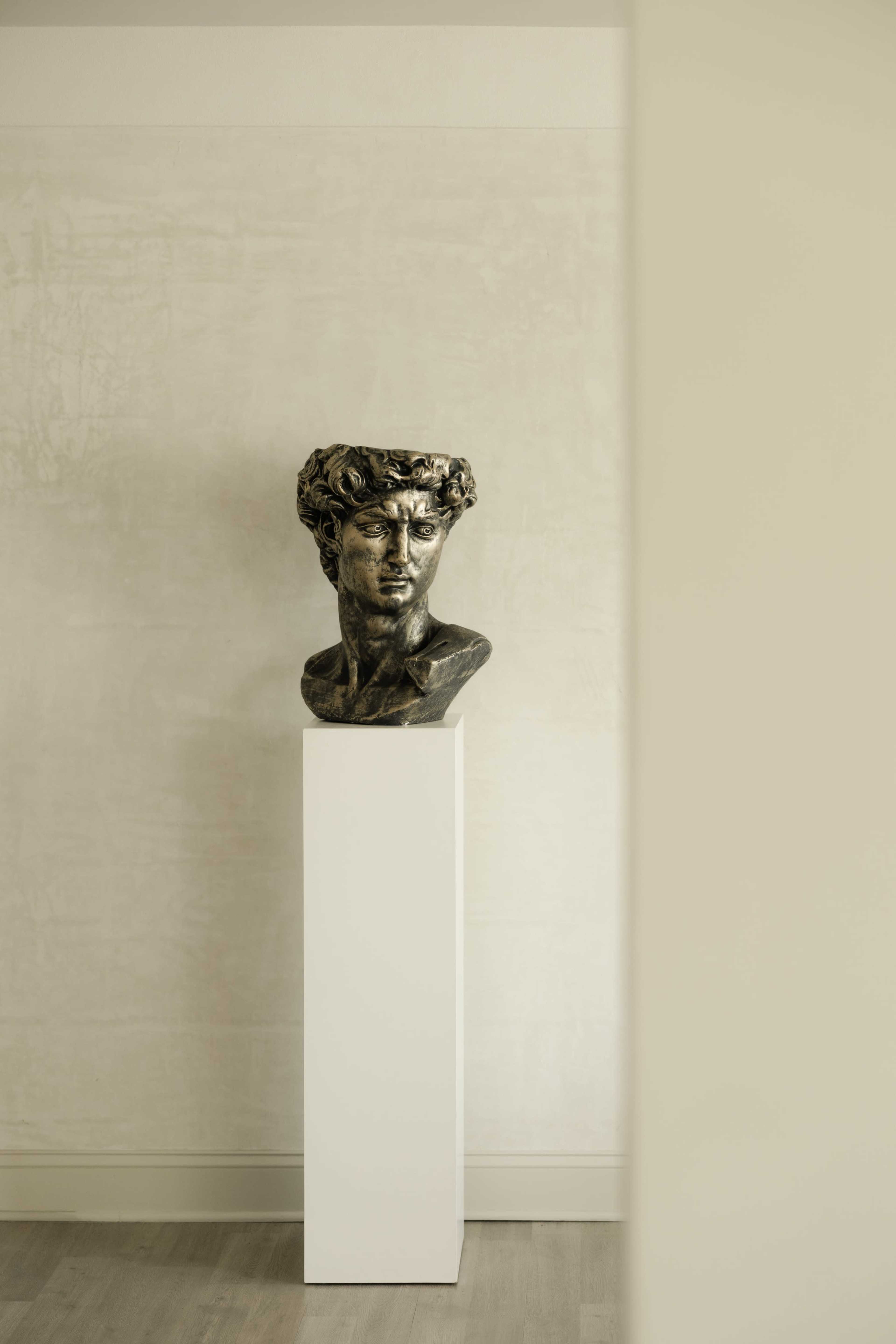 A bronze bust of a young man with curly hair is displayed on a white pedestal against a neutral wall.