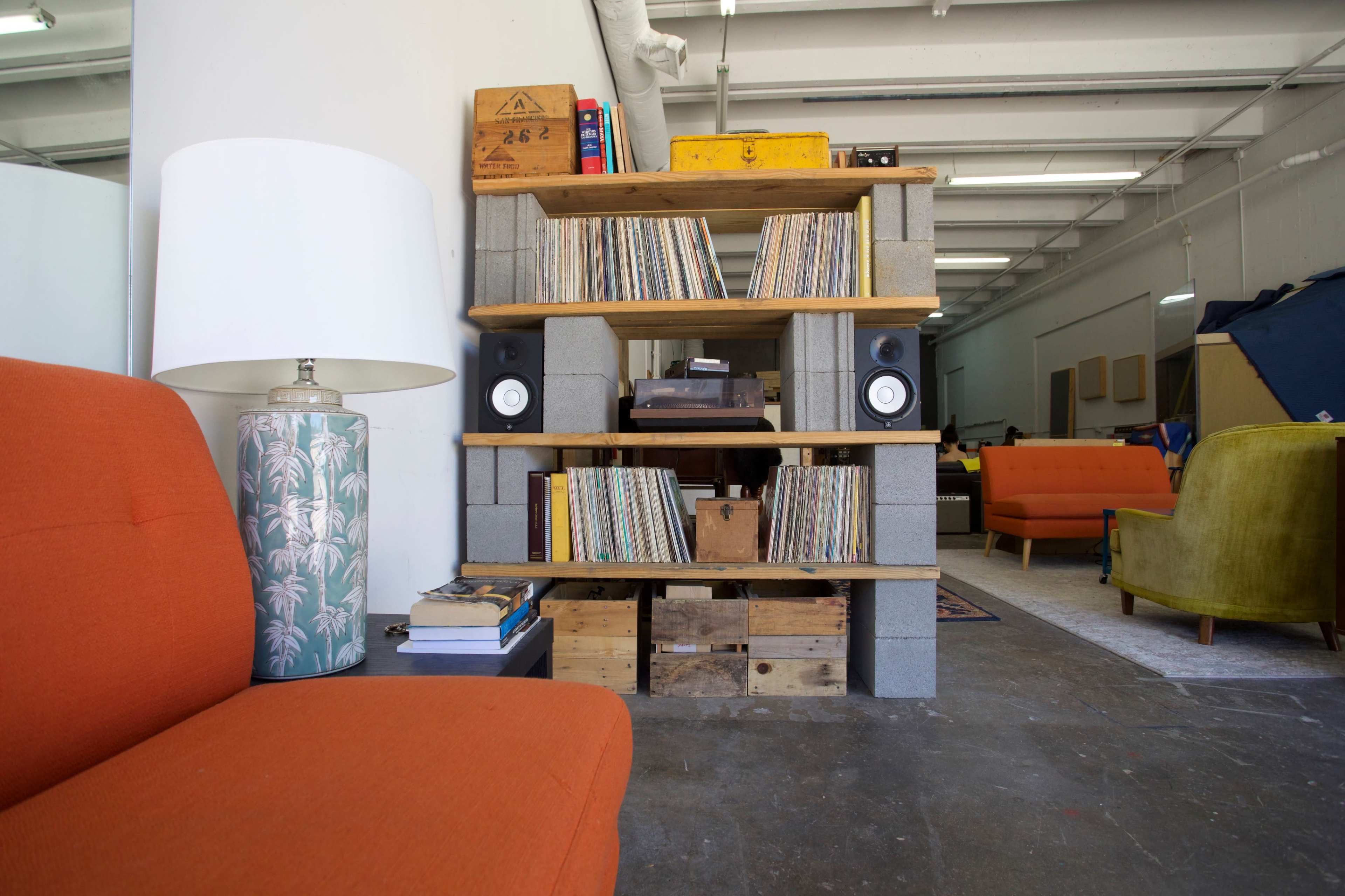 A wooden shelf made from concrete blocks holds a collection of vinyl records, flanked by speakers, with an orange couch and a lamp in the foreground.