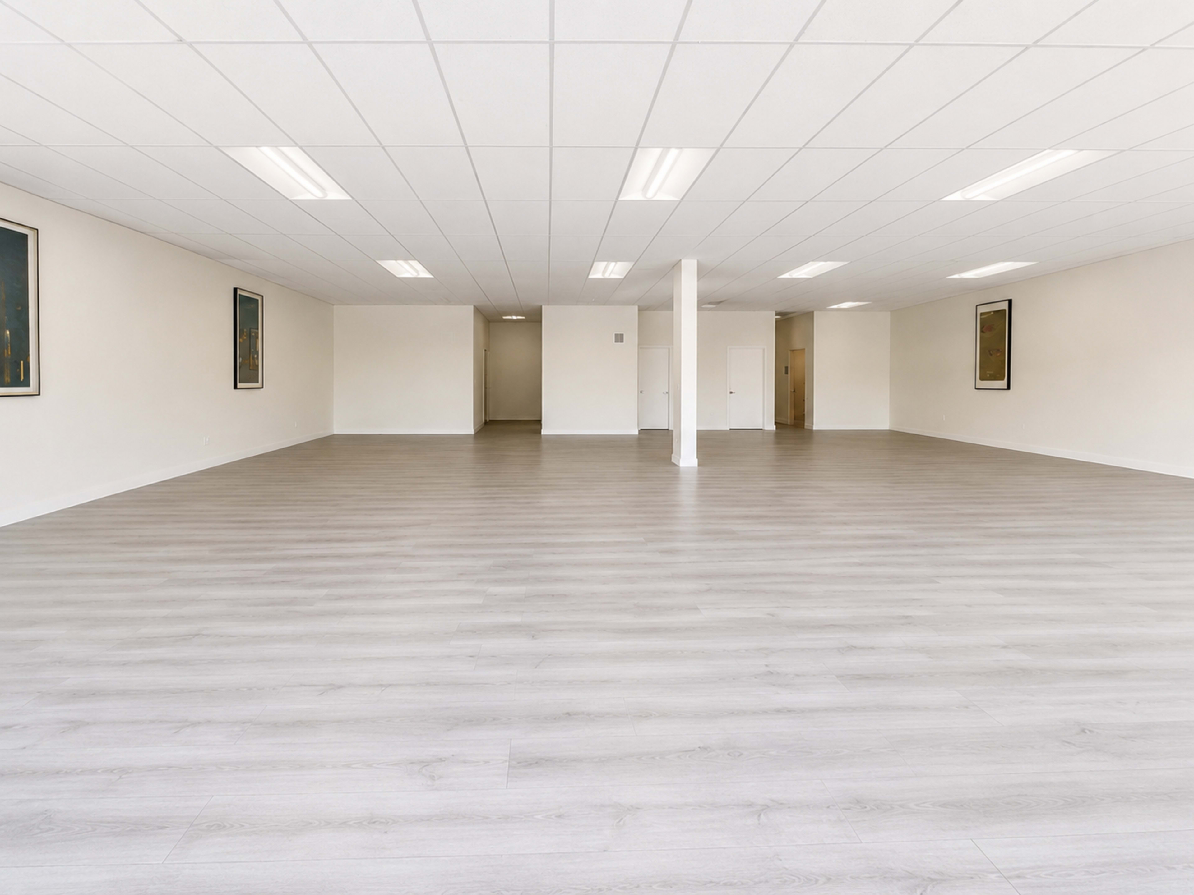 The image shows a spacious, empty room with light-colored wood flooring and minimal furnishings, featuring white walls and overhead lighting.