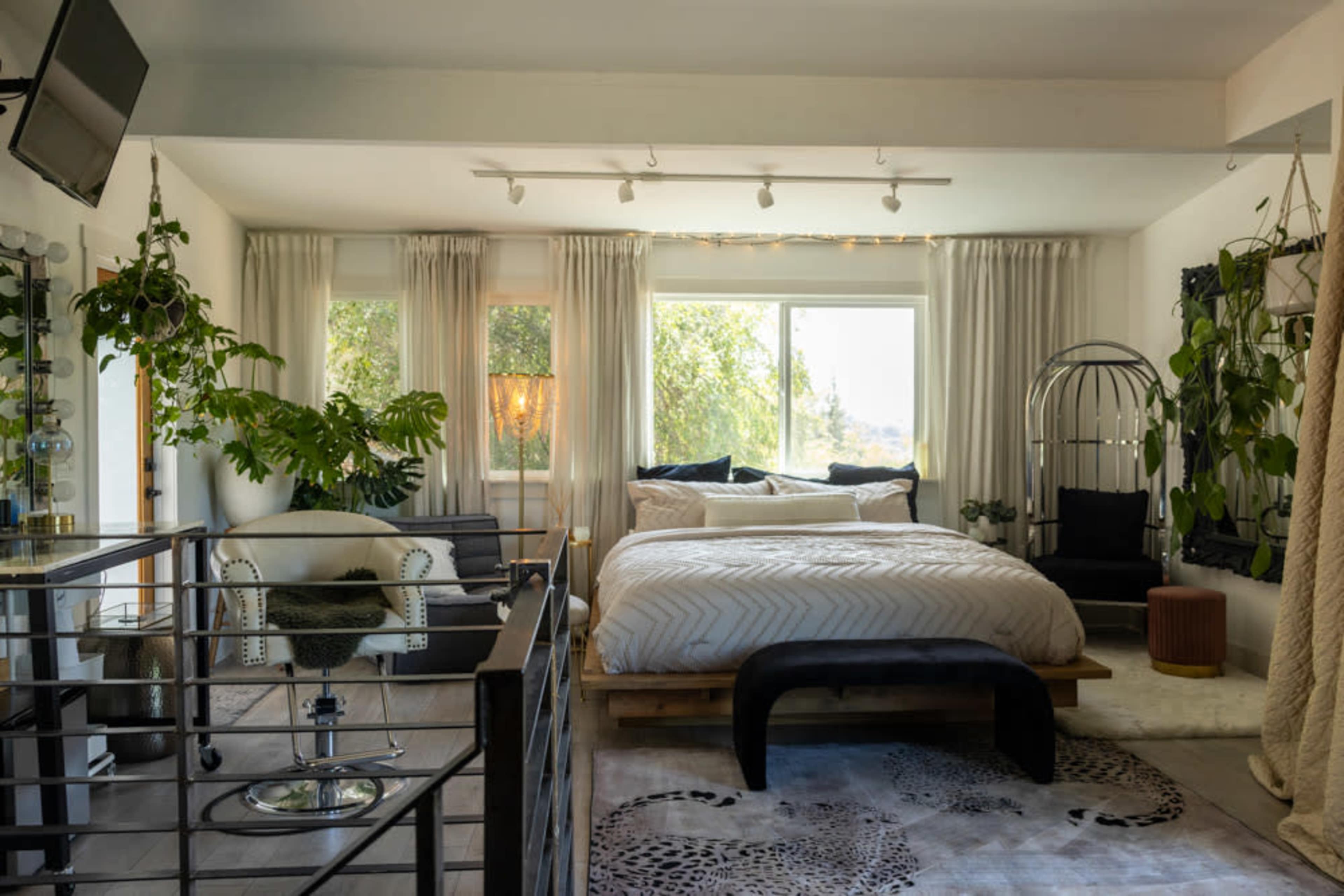 The image depicts a modern bedroom featuring a large bed, various plants, and contemporary furnishings beside a large window.