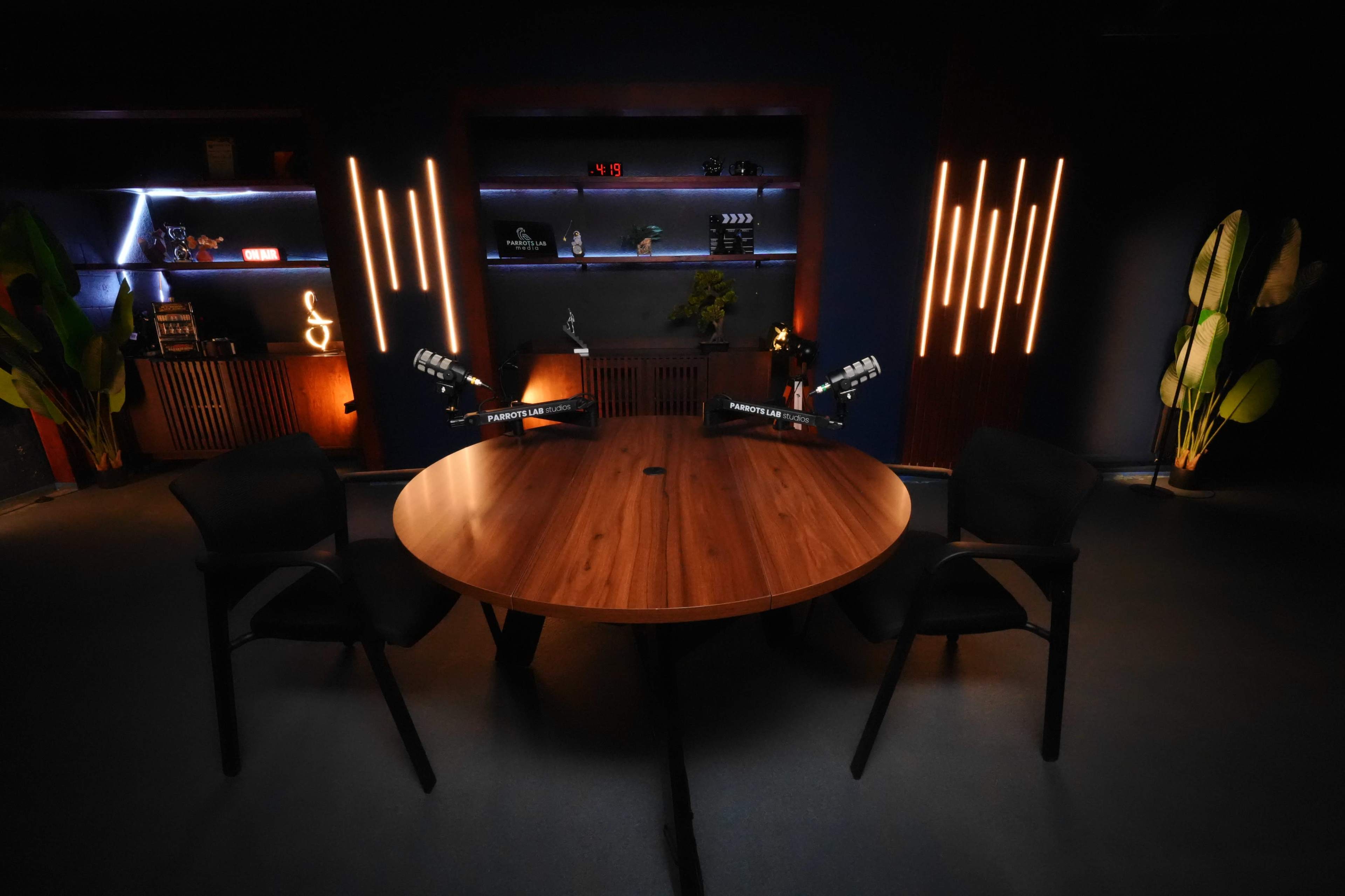 The image shows a modern podcast studio with a round wooden table and two black chairs, illuminated by ambient lighting against a dark backdrop.