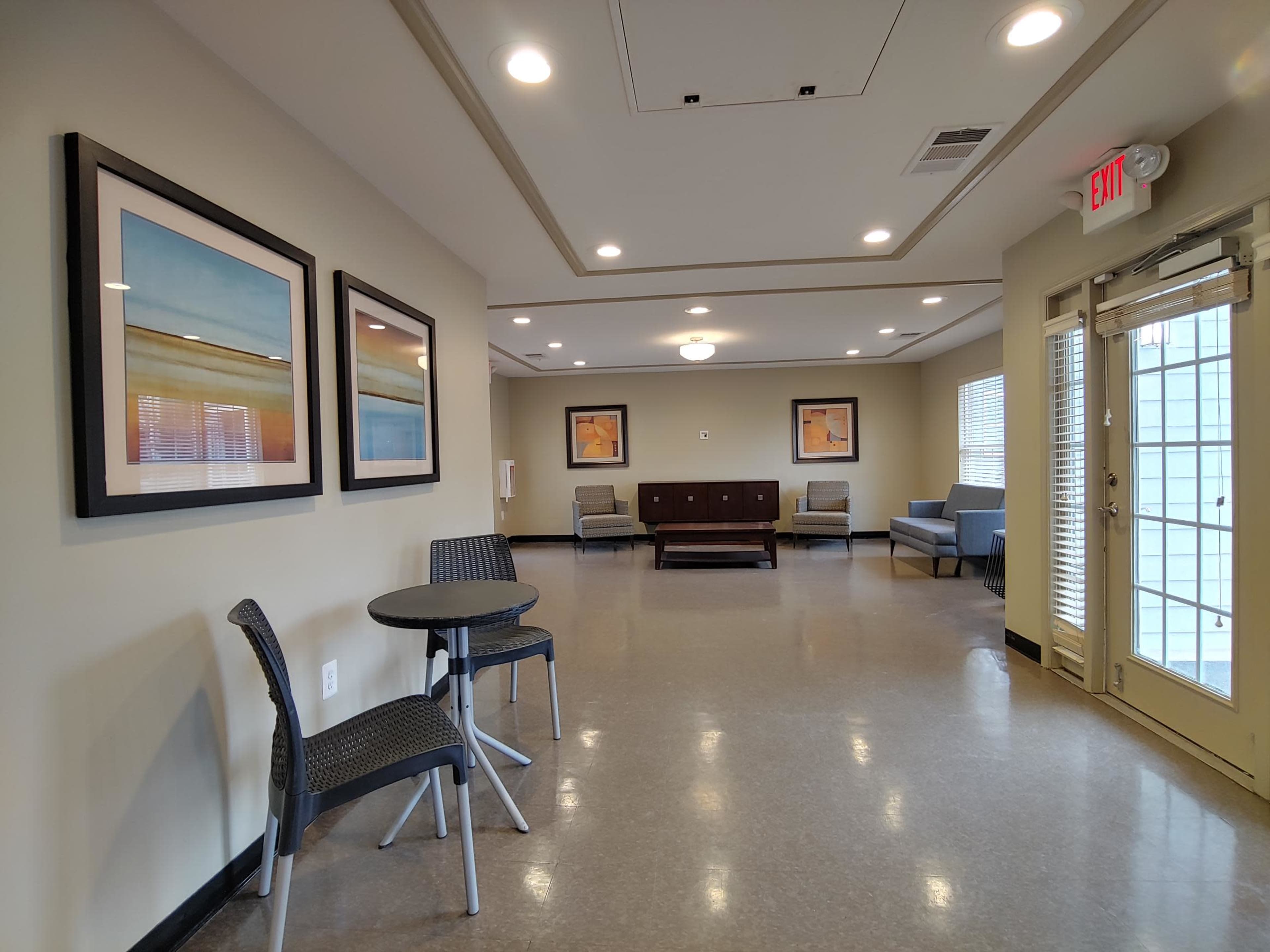 A spacious room with two framed artworks on the walls, a small round table with chairs, and seating arranged around a central wooden cabinet.