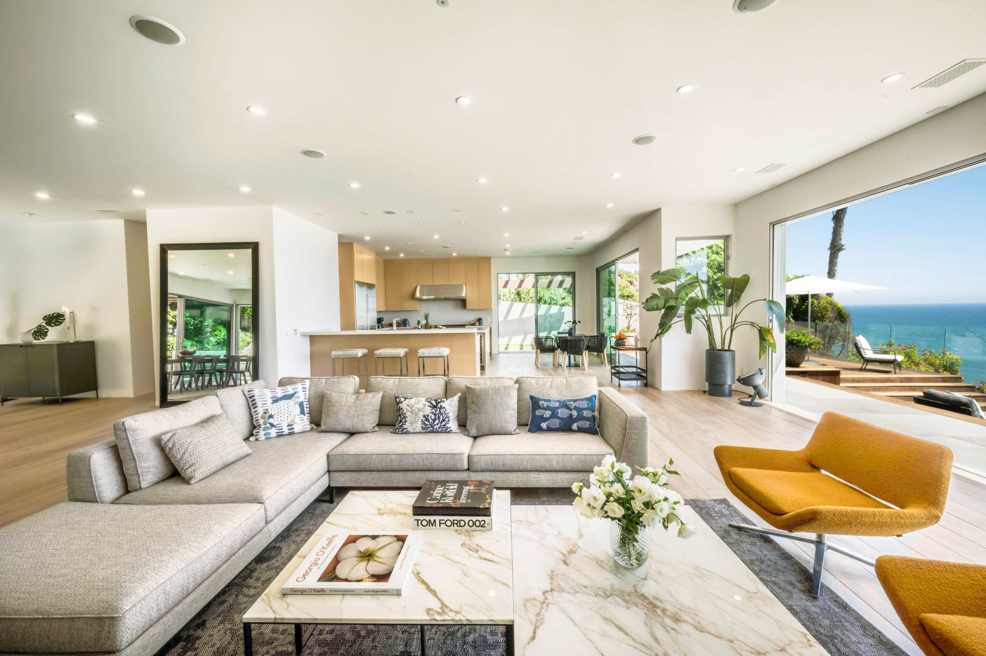 The living room features a large gray sectional sofa, a marble coffee table, and wide windows offering a view of the ocean.