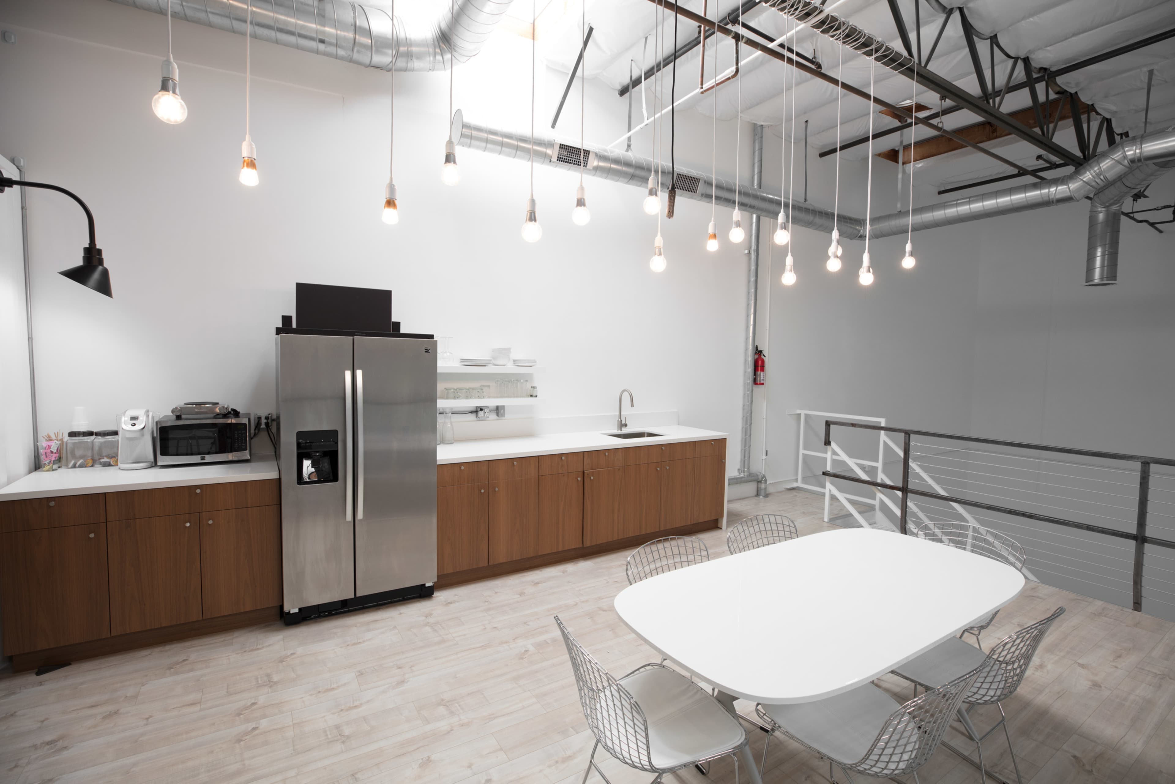 A modern kitchen area with stainless steel appliances, a long countertop, and a dining table surrounded by chairs, all set under industrial-style lighting.