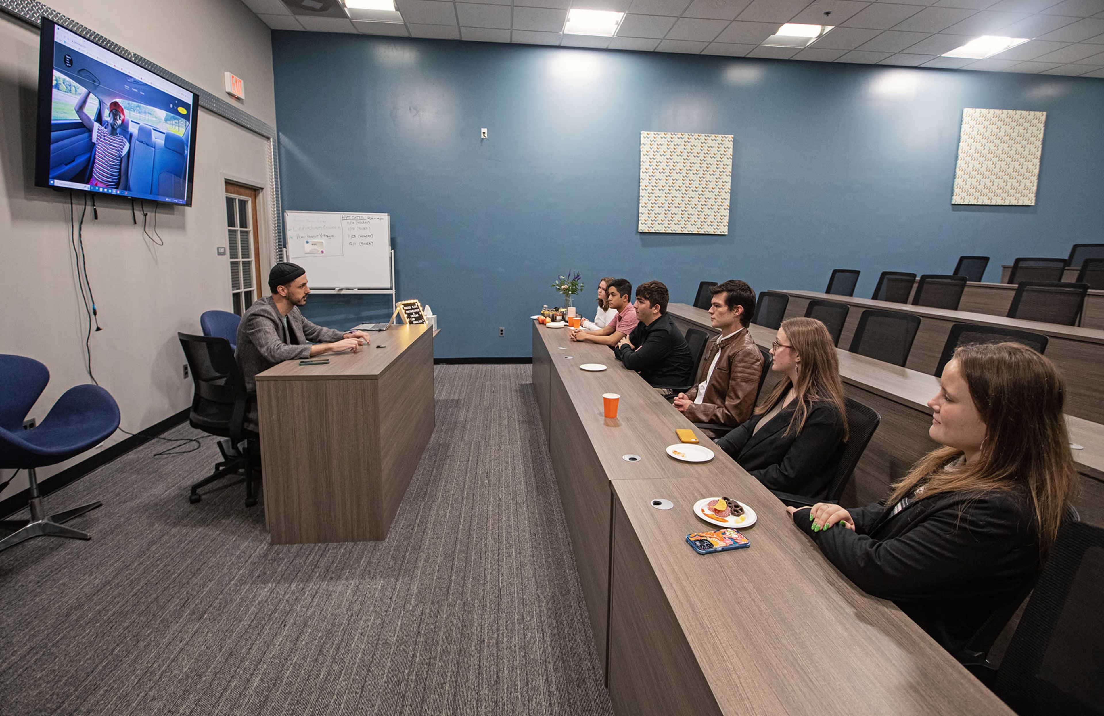 A group of seven people sits at a table facing a presenter in a classroom setting, with a screen displaying content in the background.
