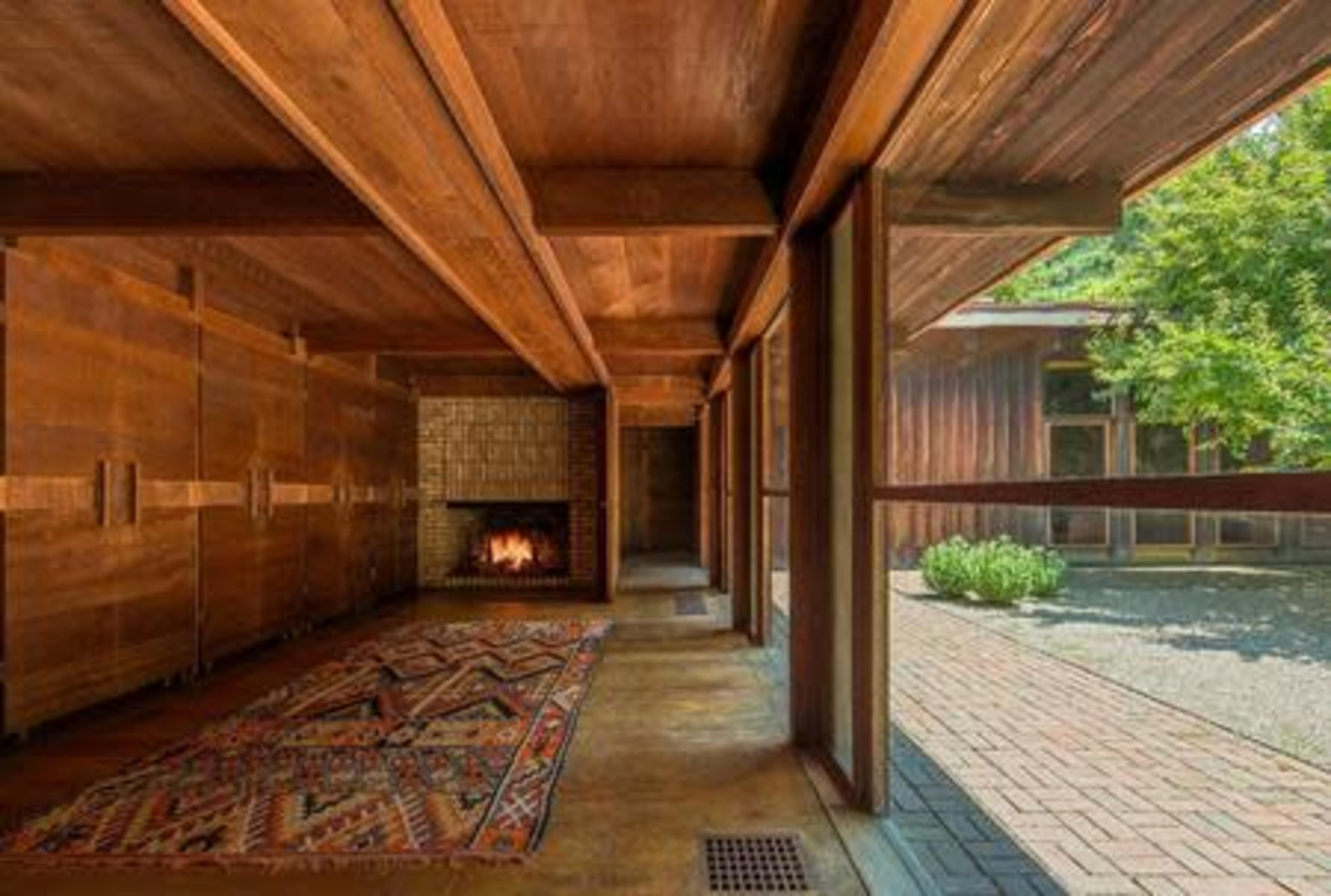 A wooden interior space featuring a fireplace and a patterned rug, with large windows opening to an outdoor area.