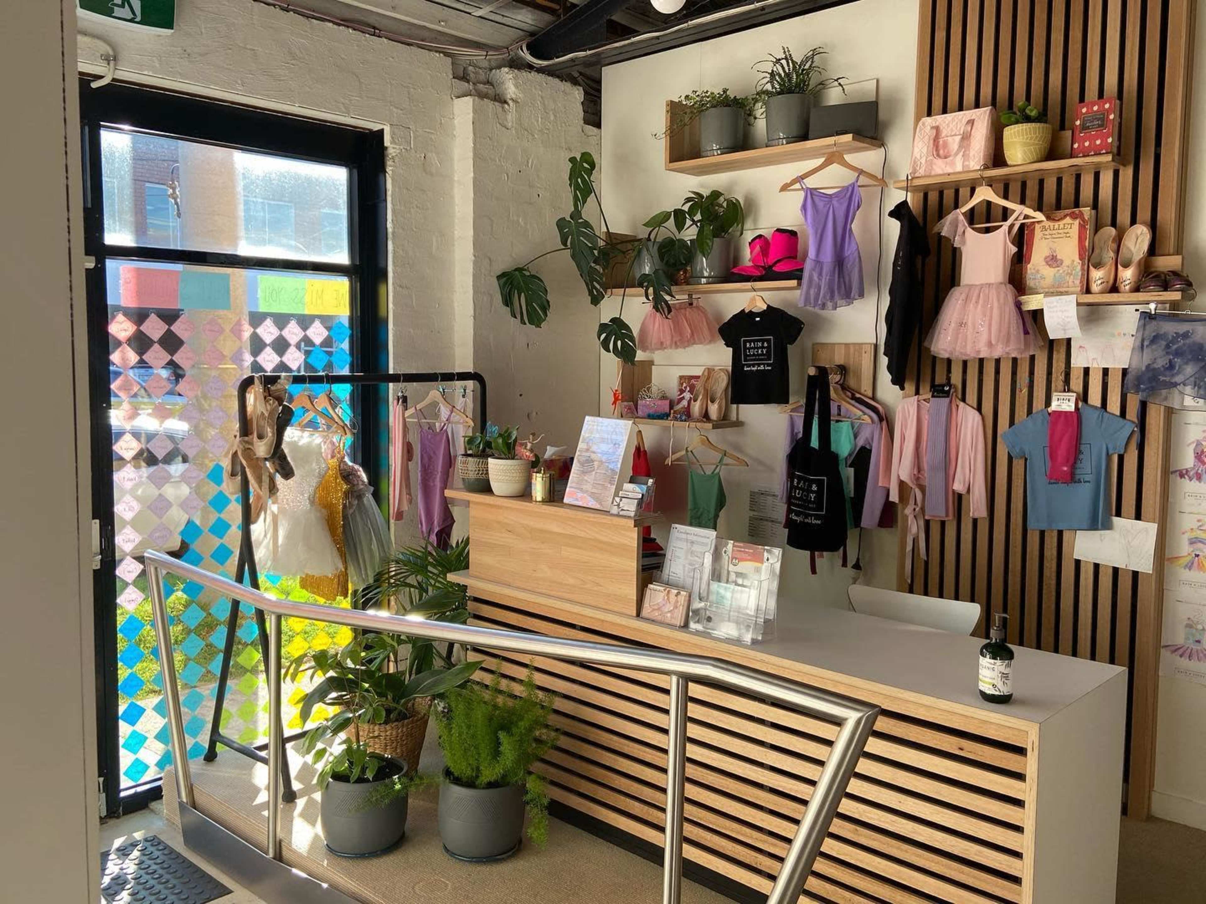 The image shows a retail space featuring a wooden counter surrounded by colorful dancewear and accessories displayed on the walls, with a vibrant glass door in the background.