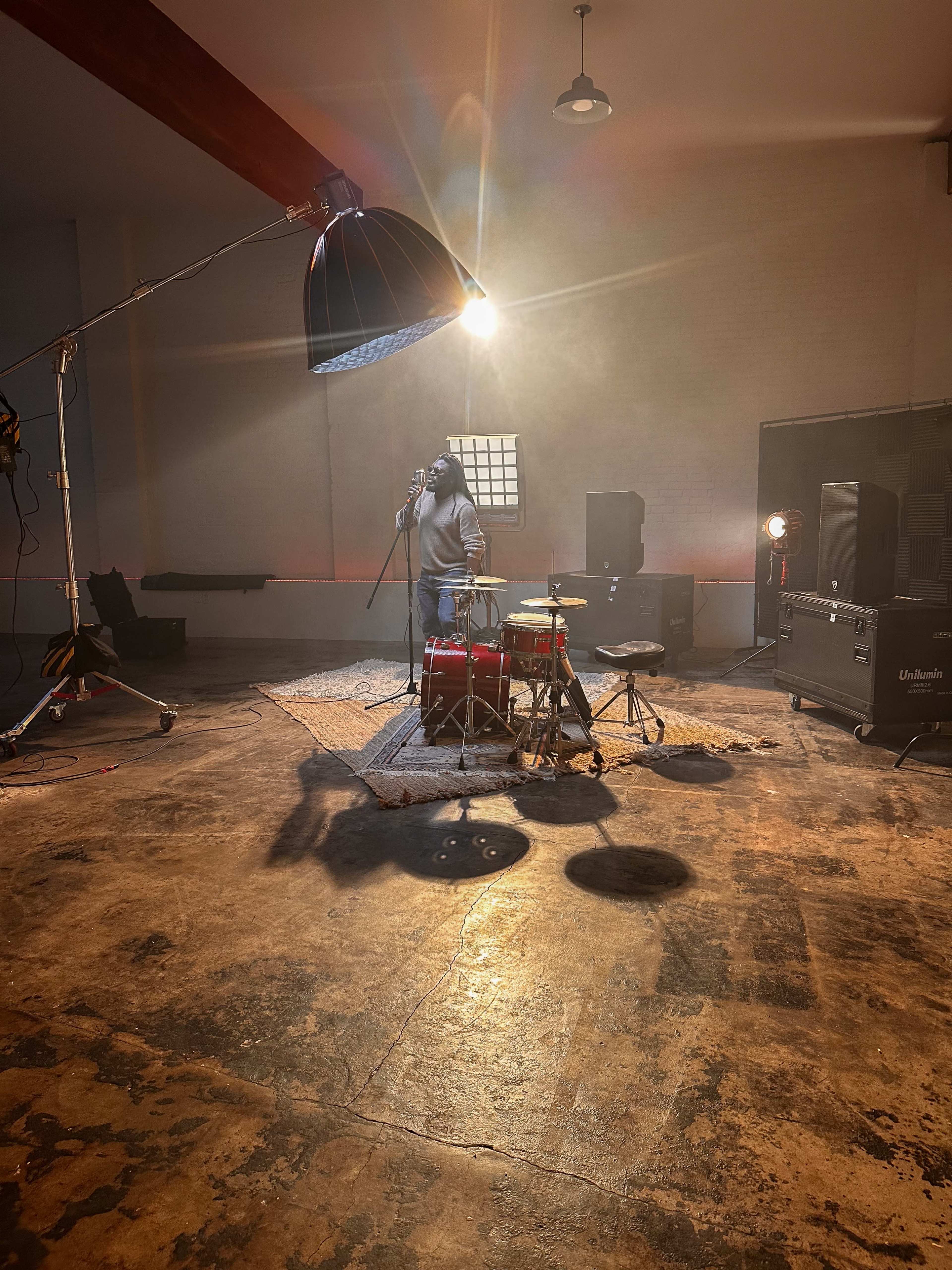 A singer stands onstage with a microphone, surrounded by a drum set and studio equipment under a single overhead light in an empty rehearsal space.