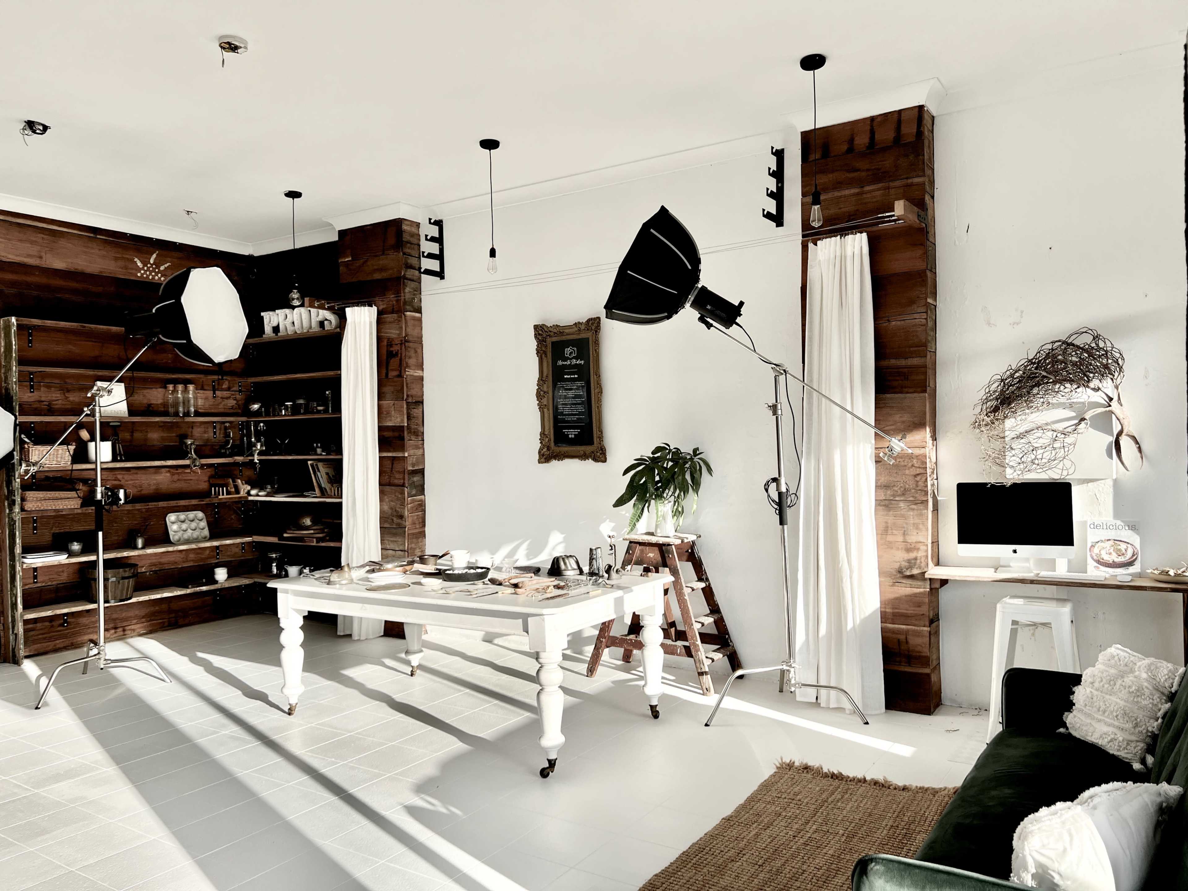The image shows a well-lit room featuring a wooden bookshelf, a large table with various items on it, and professional lighting equipment positioned around the space.