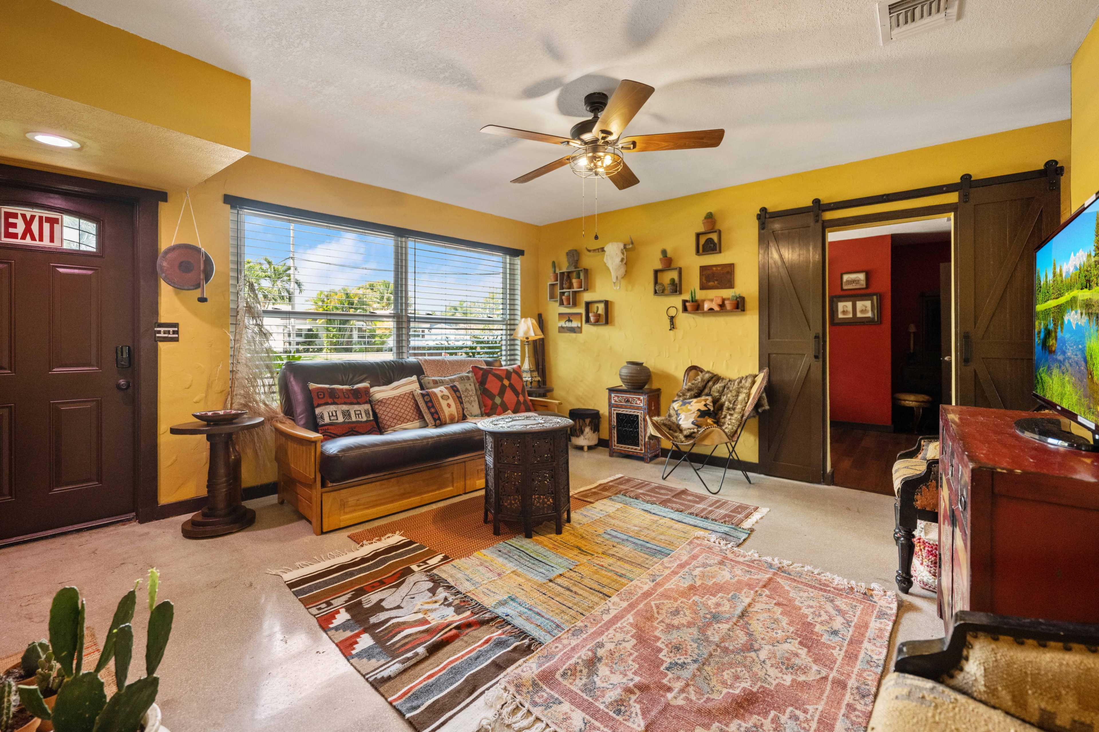 The living room features a combination of a brown leather sofa, patterned rugs, and walls painted in yellow, with a ceiling fan and decorative wall items.