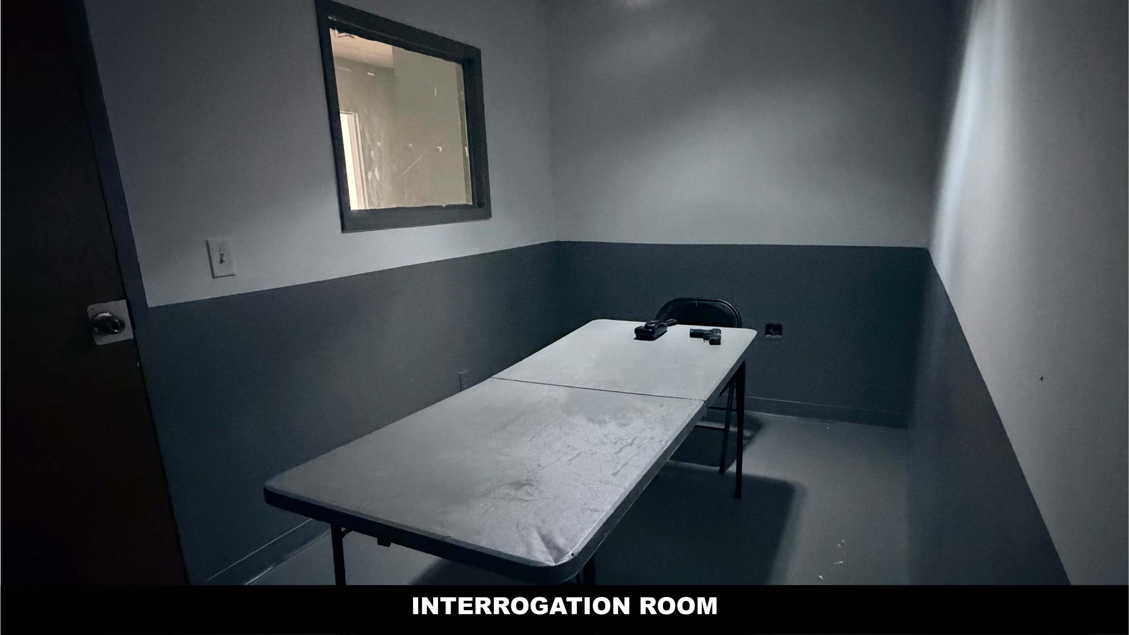 The image shows a stark interrogation room with a small table and a single chair on one side, illuminated by overhead lighting.
