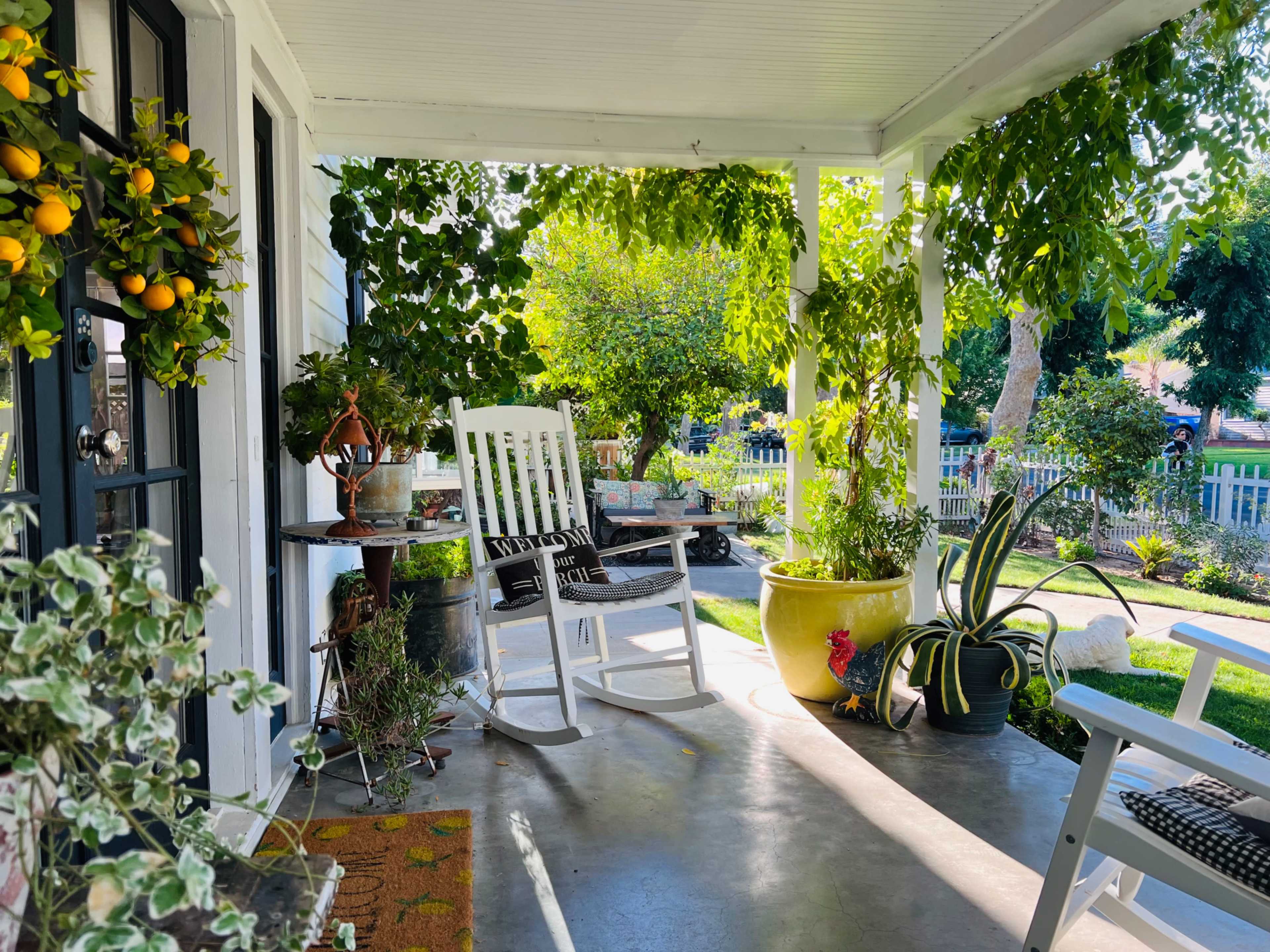 A white rocking chair sits on a porch surrounded by various potted plants and a view of a green lawn.