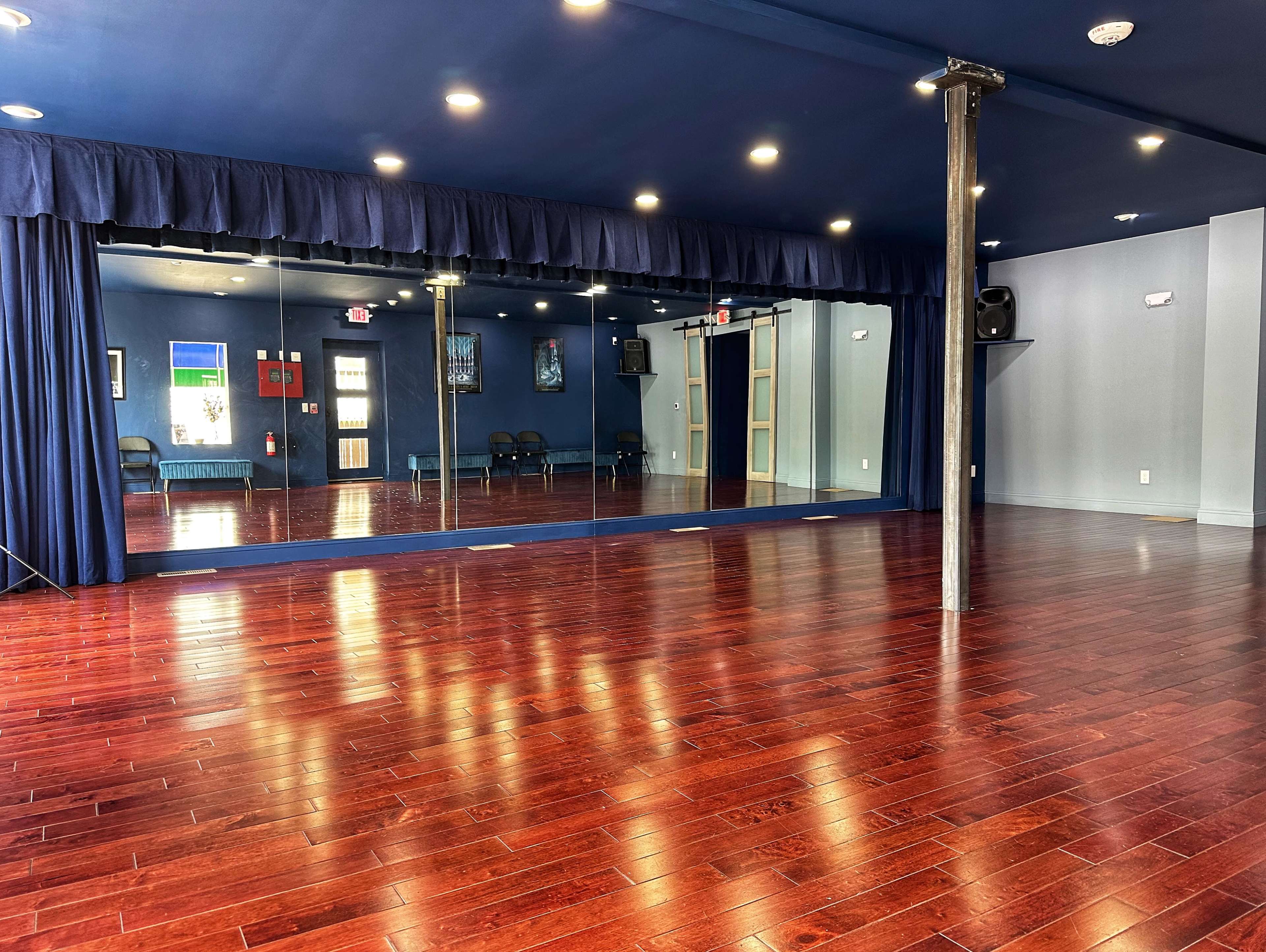 The image shows a dance studio with a wooden floor, a large mirror along one wall, and blue walls with ceiling lights.