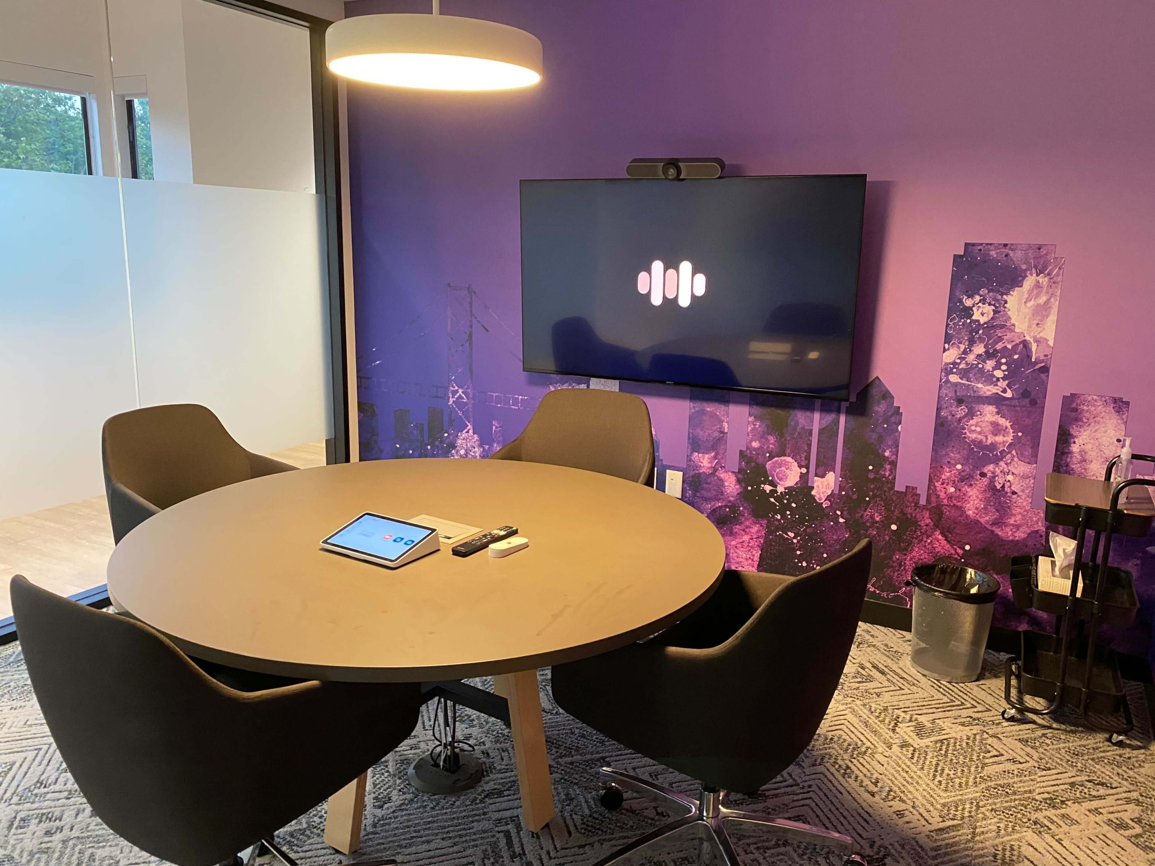 A modern conference room features a round table with four chairs, a large television on the wall displaying a logo, and an iPad and remote controls on the table.