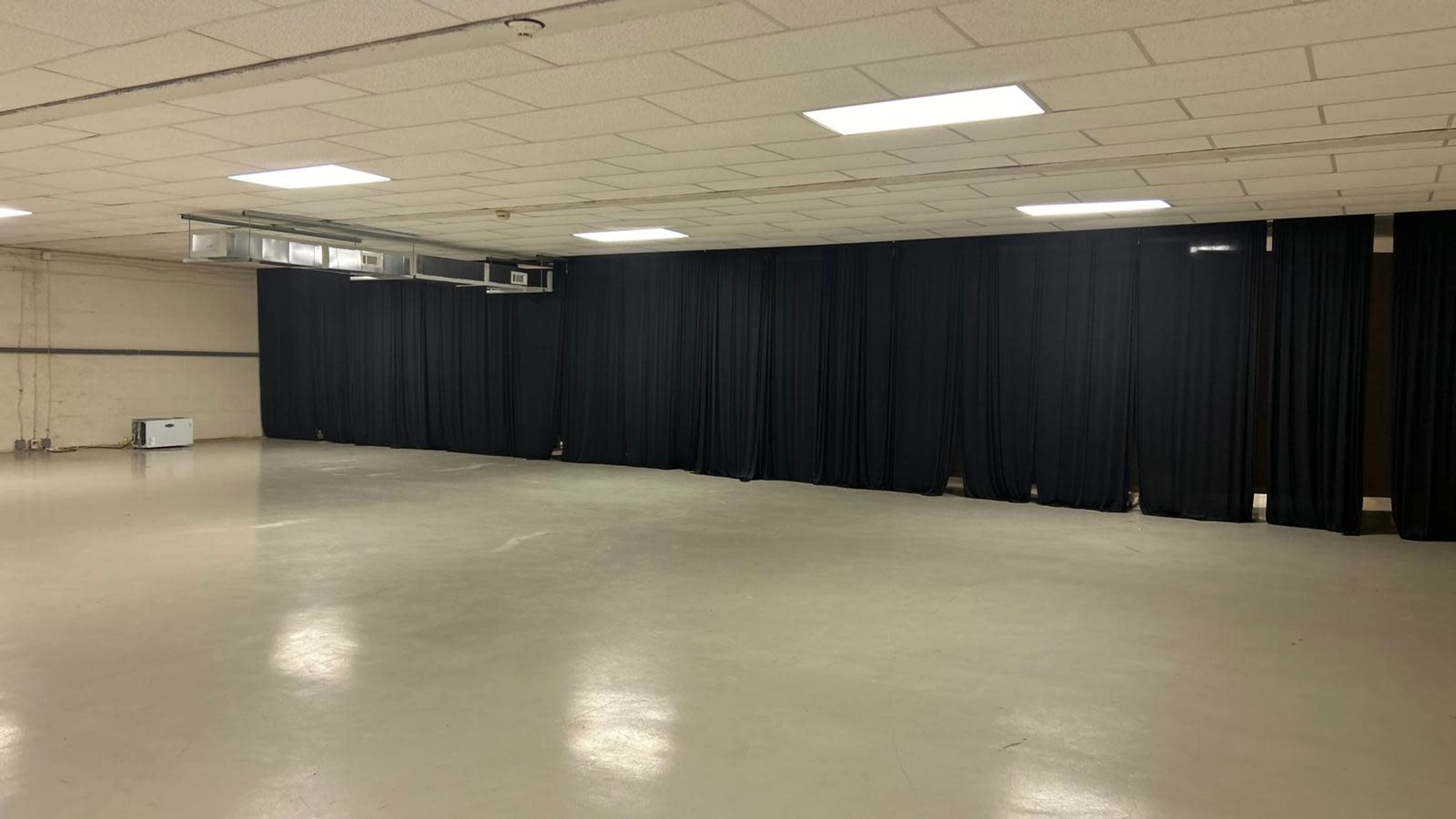 The image shows a large, empty room with a gray floor and black curtains lining one wall.