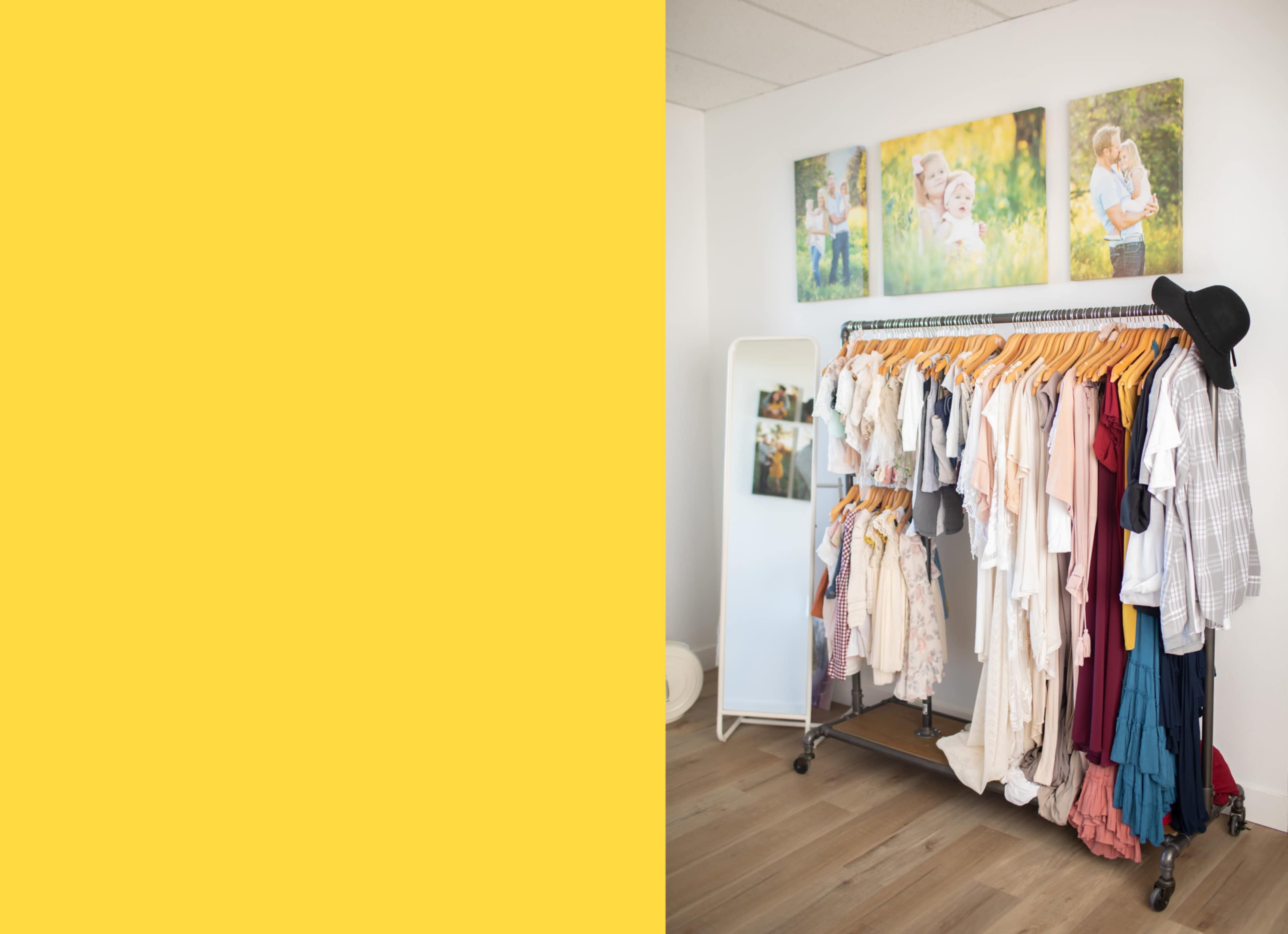 A clothing rack filled with various garments is positioned in a well-lit room featuring framed photographs on the wall.