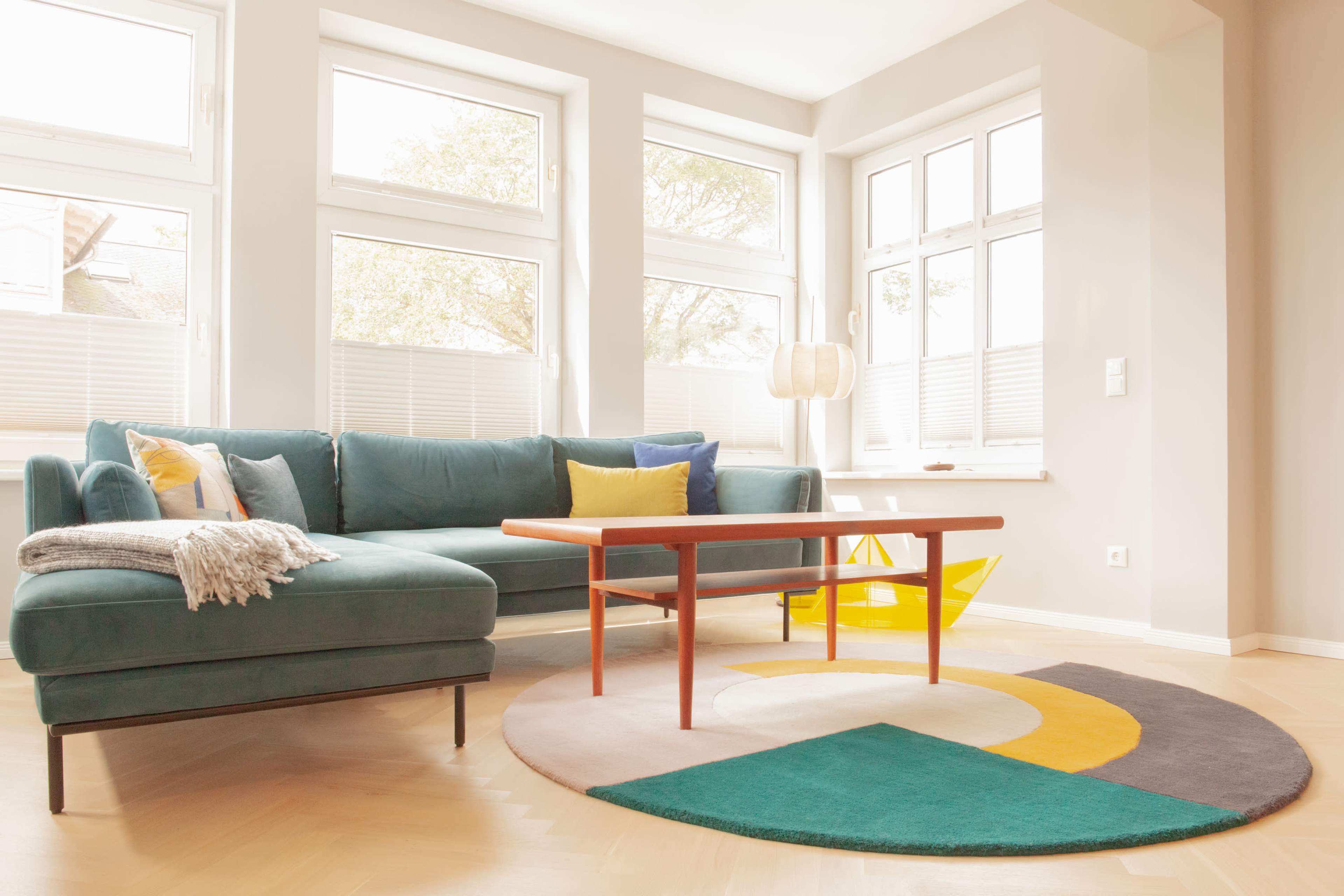A modern living room featuring a teal sectional sofa, a wooden coffee table, and a circular rug with geometric patterns, illuminated by natural light from the surrounding windows.