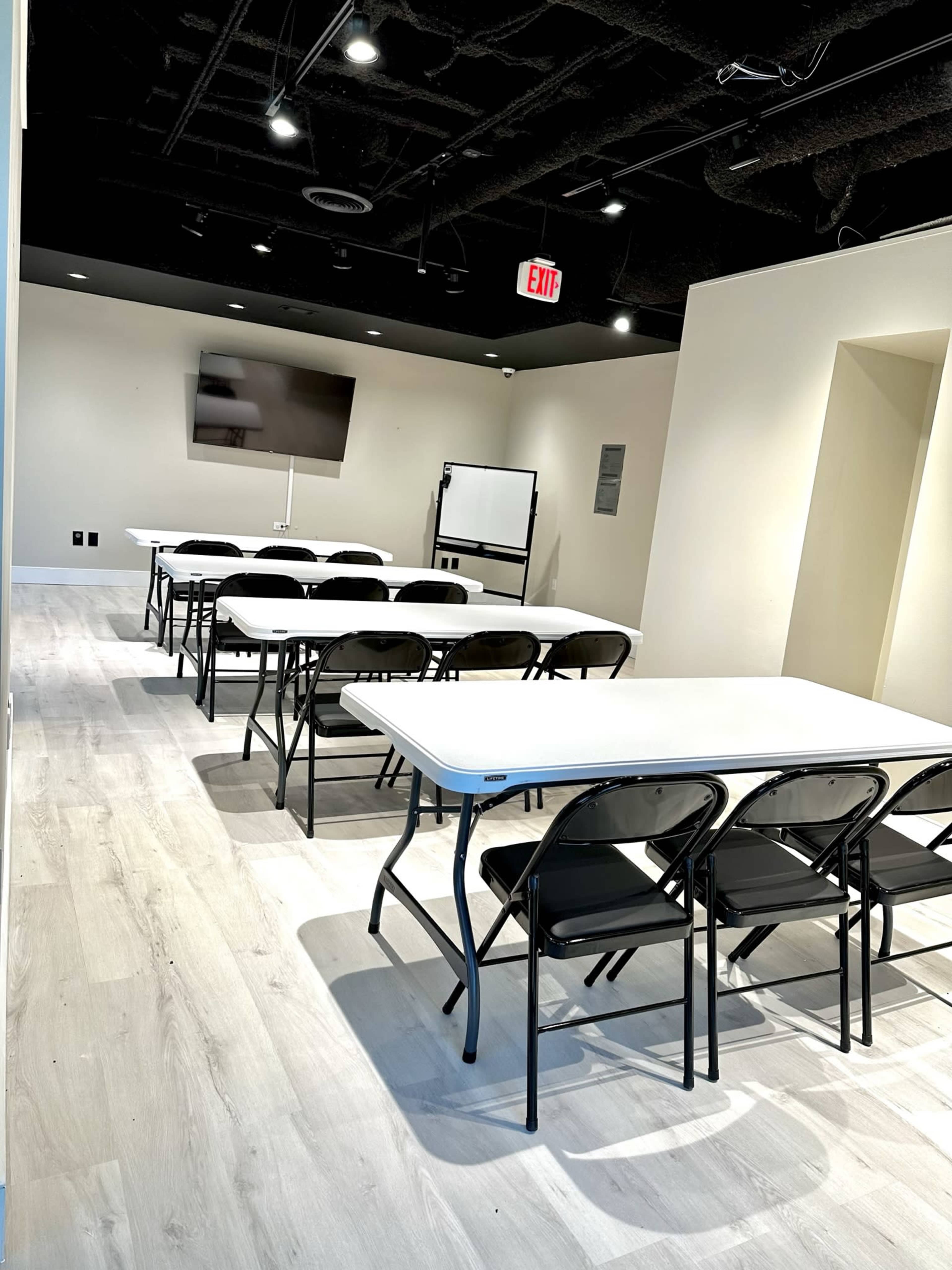 A series of rows of folding tables and chairs set up in a brightly lit meeting room, with a television screen and a whiteboard at the front.