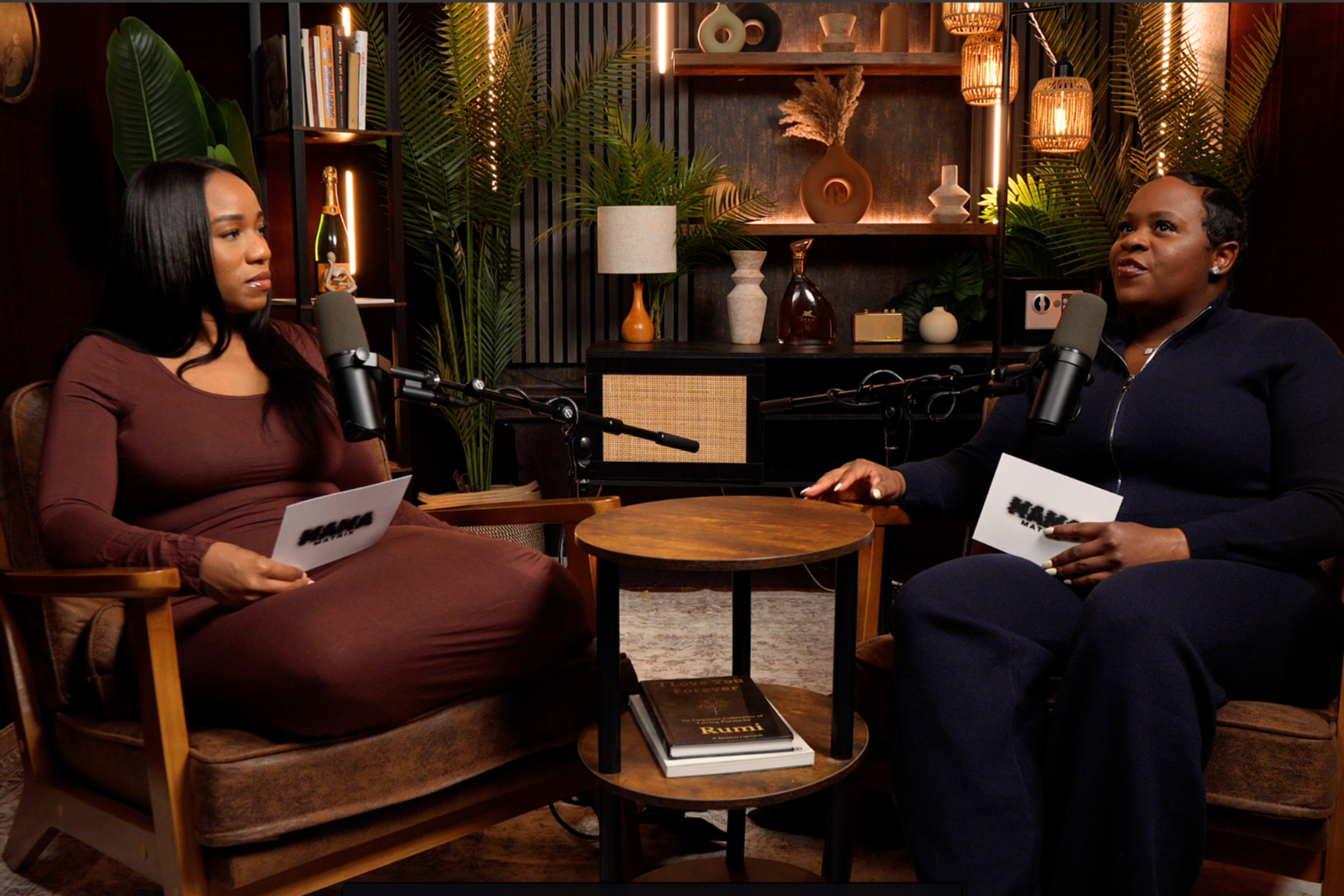 Two women sit in armchairs with microphones, engaged in conversation in a well-decorated studio setting.