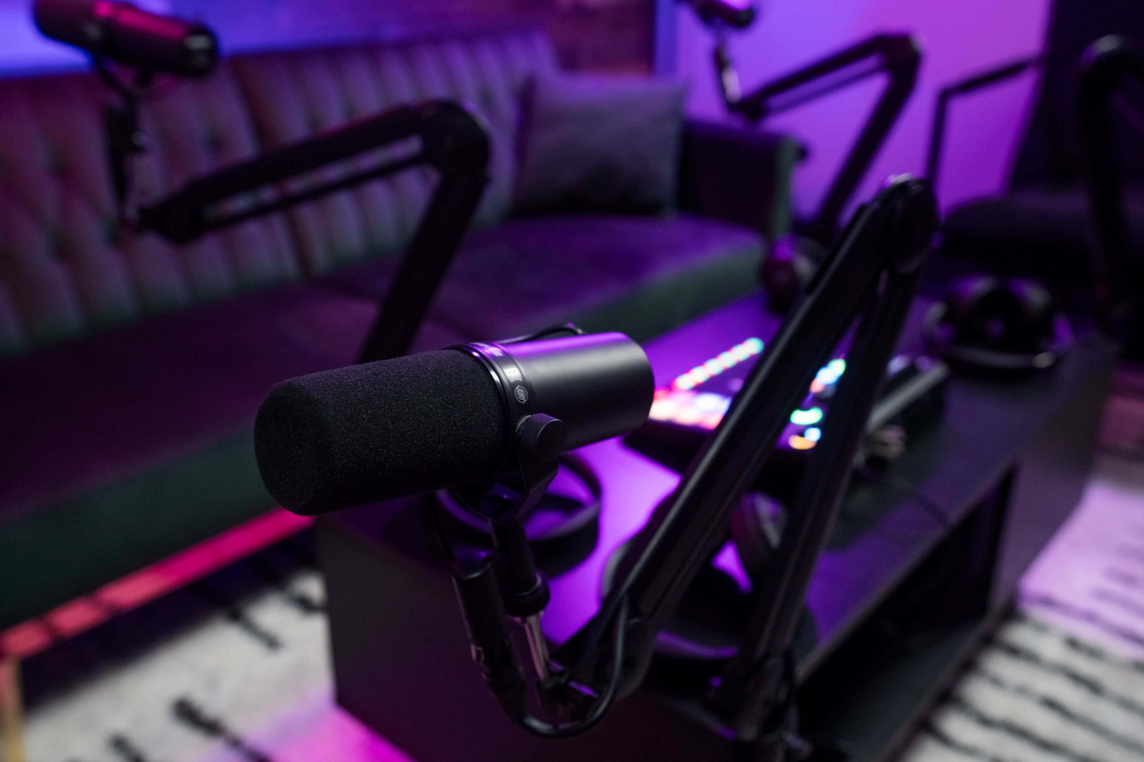 A black microphone is positioned on a stand in a podcasting studio, with a green couch and colorful soundboard visible in the background.